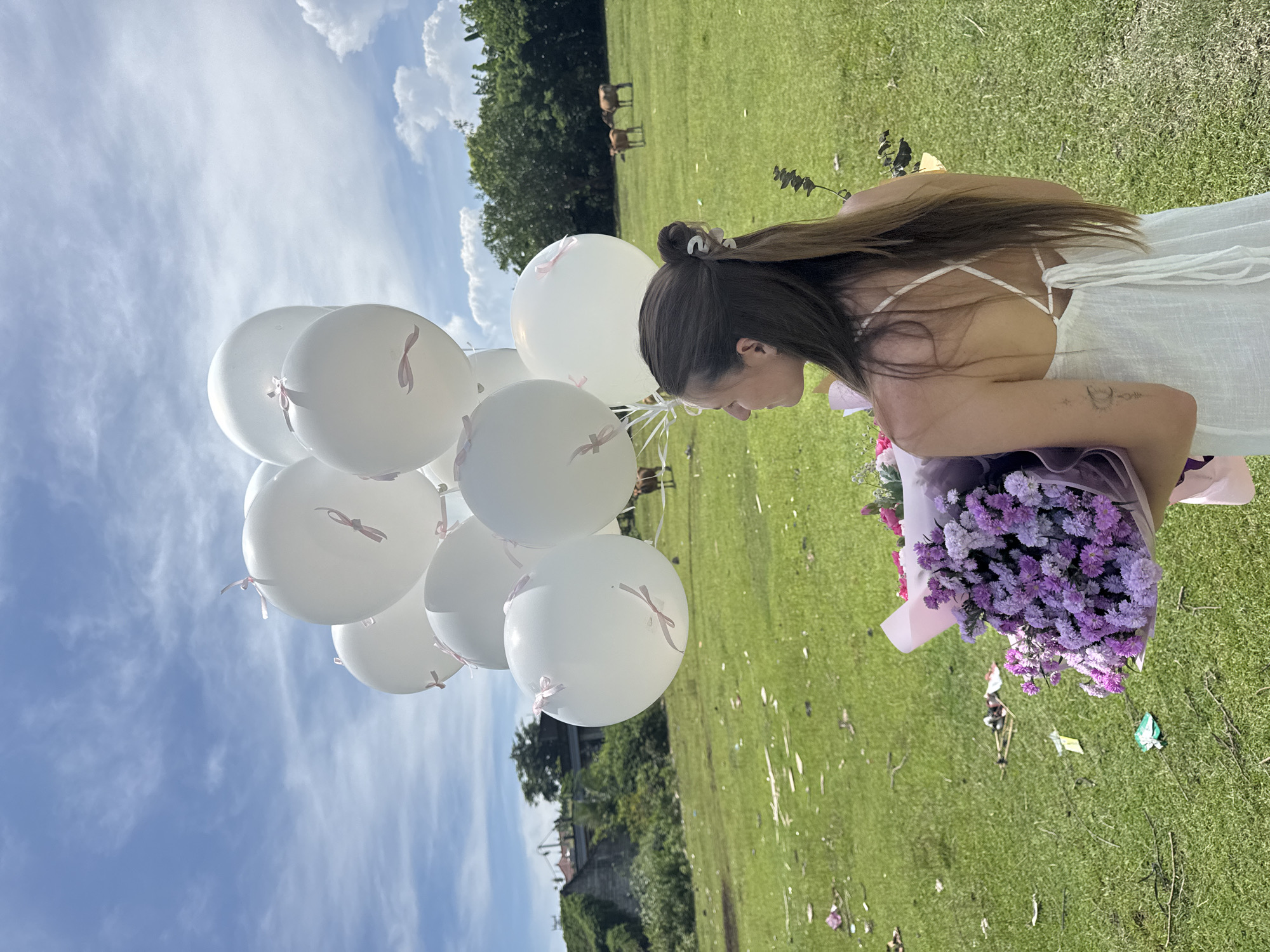 A medium shot depicts a woman in a white dress outdoors, holding a bouquet of purple flowers and a bunch of white balloons. The background shows a green field with sparse trees and a blue sky with scattered clouds. Horses are faintly visible in the distance. The image is oriented sideways.