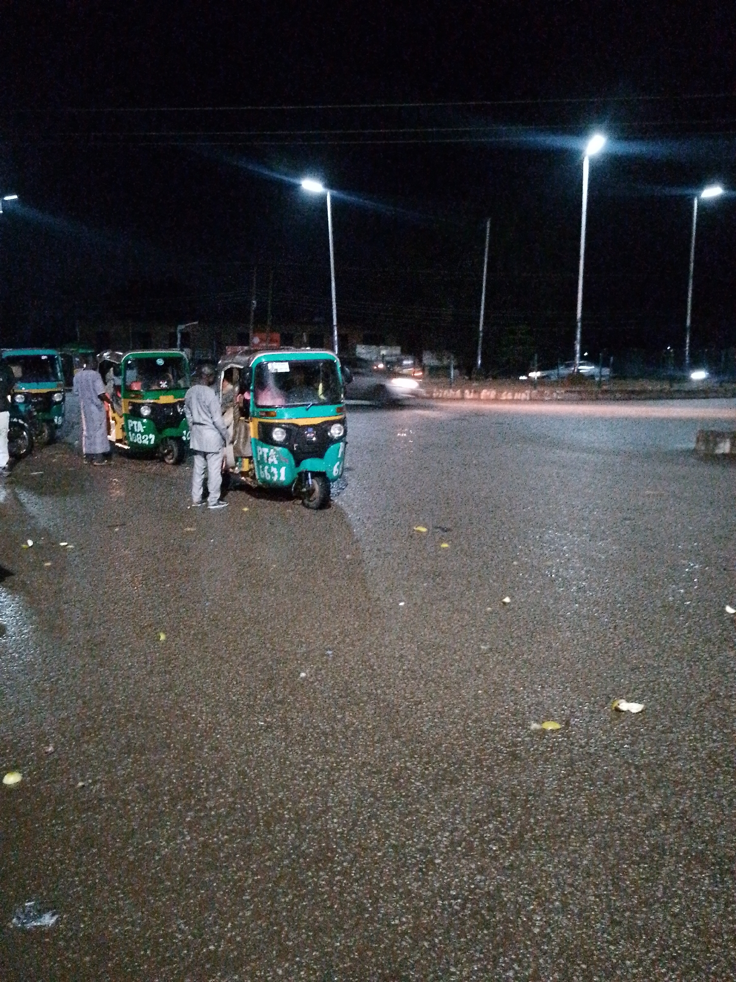 This image depicts a nighttime scene at what appears to be a transportation hub or waiting area in Bukuru, Nigeria. Several three-wheeled auto-rickshaws, commonly known as Keke Napep or Marwa, are parked on a wet, asphalt surface. The vehicles are predominantly green with yellow and black accents. Some have visible license plates or markings, such as "PTA 10827" and "PTA 6631".
At least three individuals are visible. One person, wearing a light-colored traditional outfit, stands near the first Keke. Another person, in a similar light-colored garment, stands between two Keke, facing away from the camera. A third individual, dressed in a light grey suit, stands behind one of the Keke, also facing away. The lighting in the scene is provided by streetlights, which cast long shadows and create a somewhat hazy atmosphere. In the background, distant lights suggest the presence of buildings and moving vehicles on a road. The ground is speckled with small, scattered yellow and white debris, possibly fallen leaves or trash, and appears damp, indicating recent rain or ongoing moisture. The overall impression is of a quiet, possibly a resting or waiting period for these local transportation vehicles and their operators during the night.