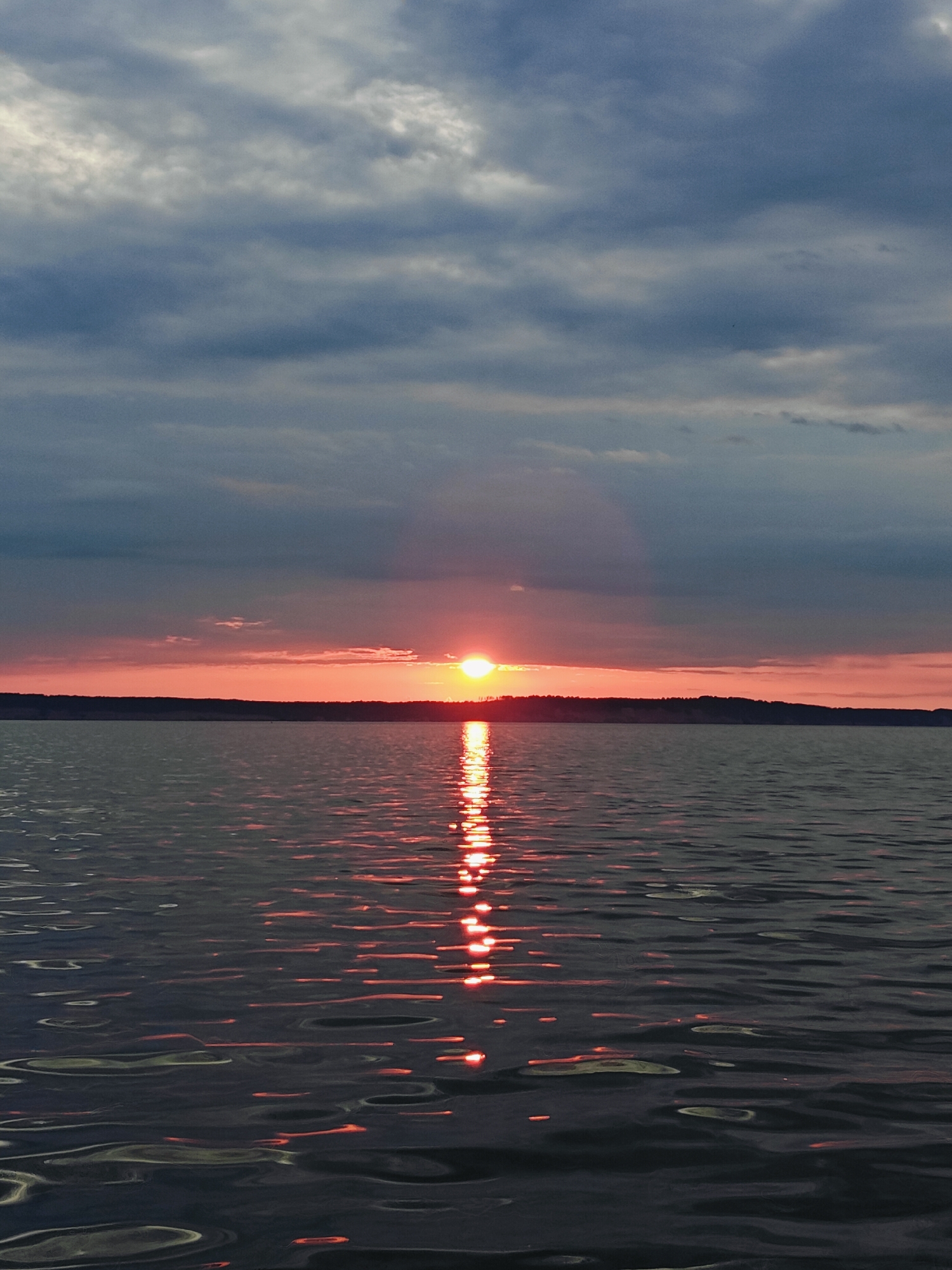 This is a vertical, eye-level, outdoor, full-shot photograph of a sunset over a body of water. The sun is setting on the horizon, casting a bright orange glow that blends into a soft pink and then into the dark gray clouds above. The sun's reflection is visible on the water's surface as a shimmering, golden path. The water itself is dark and appears to be rippling slightly. The distant shore is silhouetted against the colorful sky and is covered with trees. The overall mood of the image is tranquil and serene. This scene is characteristic of the Kama River in Chaykovsky, Russia, where such sunsets are common. There are no people or specific objects visible in the image other than the natural landscape.