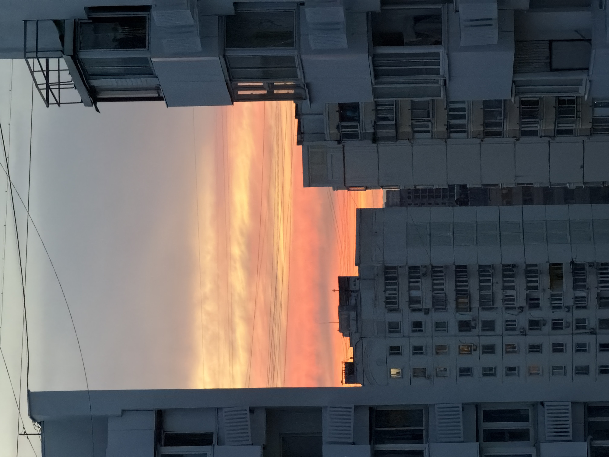 The image depicts a sunset in Khimki, Russia, framed by two buildings. The sky shows a gradient of colors ranging from gray to orange. The buildings are white and appear to be residential. The structures have many windows, with some air conditioning units also visible. There are also power lines visible on the left side of the image.