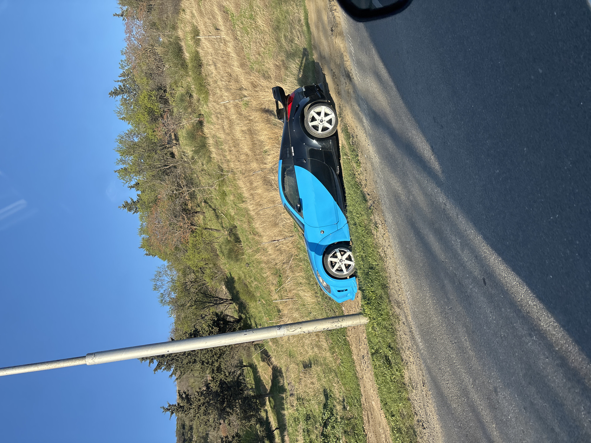 A two-toned, black and blue, sports car, parked on the grass next to a road, faces uphill. The wheels are silver with a six-spoke design. The car is parked near a field of yellowing grass and trees. A grey metal pole runs diagonally across the lower left of the frame against a blue sky. A section of asphalt road fills the right side of the frame. The location may be near Tbilisi, Georgia.
