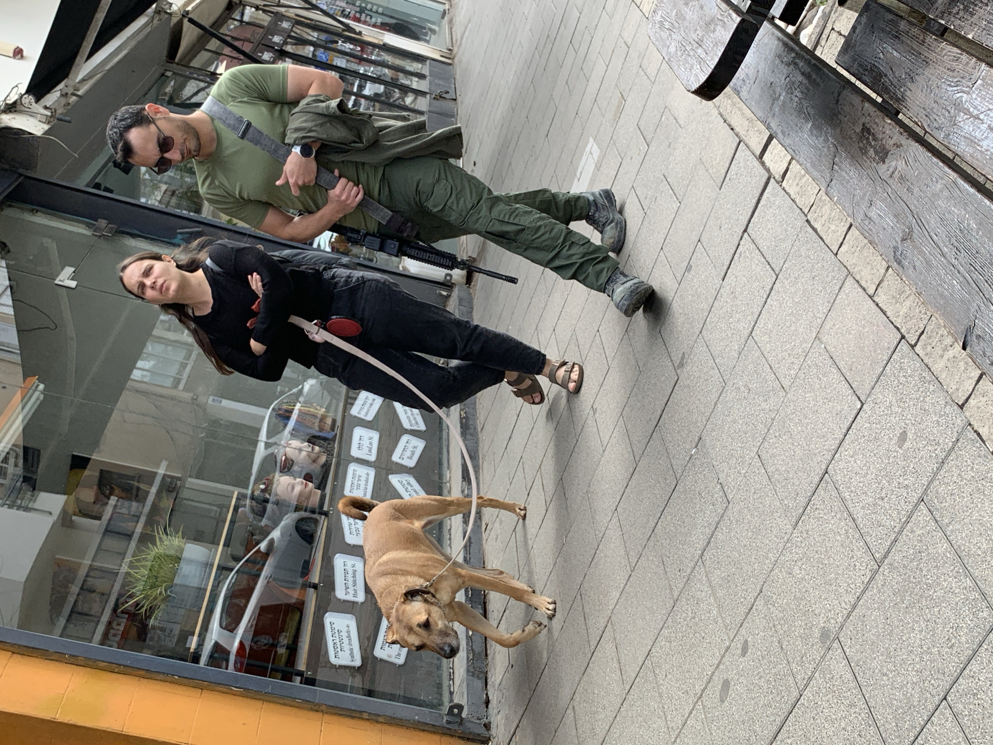 Outside a shop in Tel Aviv, Israel, a man and woman walk a light brown dog along a sidewalk. The man wears an olive t-shirt, cargo pants, sunglasses, and a strap across his chest. He carries what appears to be a rifle. The woman wears a black shirt and pants, with sandals. The shop's window displays Hebrew writing and the reflection of a car. A bench sits on the sidewalk. The sidewalk is paved with grey stone tiles.