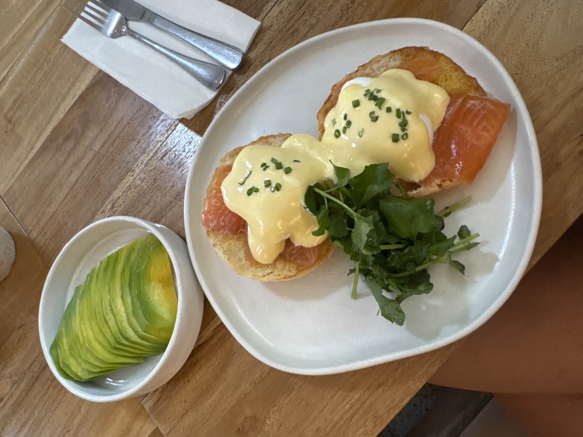 A top-down shot features a plate of Eggs Benedict with salmon and a side of sliced avocado on a wooden table. The Eggs Benedict consists of two halves of an English muffin, each topped with salmon slices, a poached egg, and hollandaise sauce garnished with chives. A small clump of green leaves is between the two halves. A small white bowl filled with thinly sliced avocado is to the left of the plate. A knife and fork rest on a white napkin in the upper-left corner. The location is North Kuta, Indonesia.
