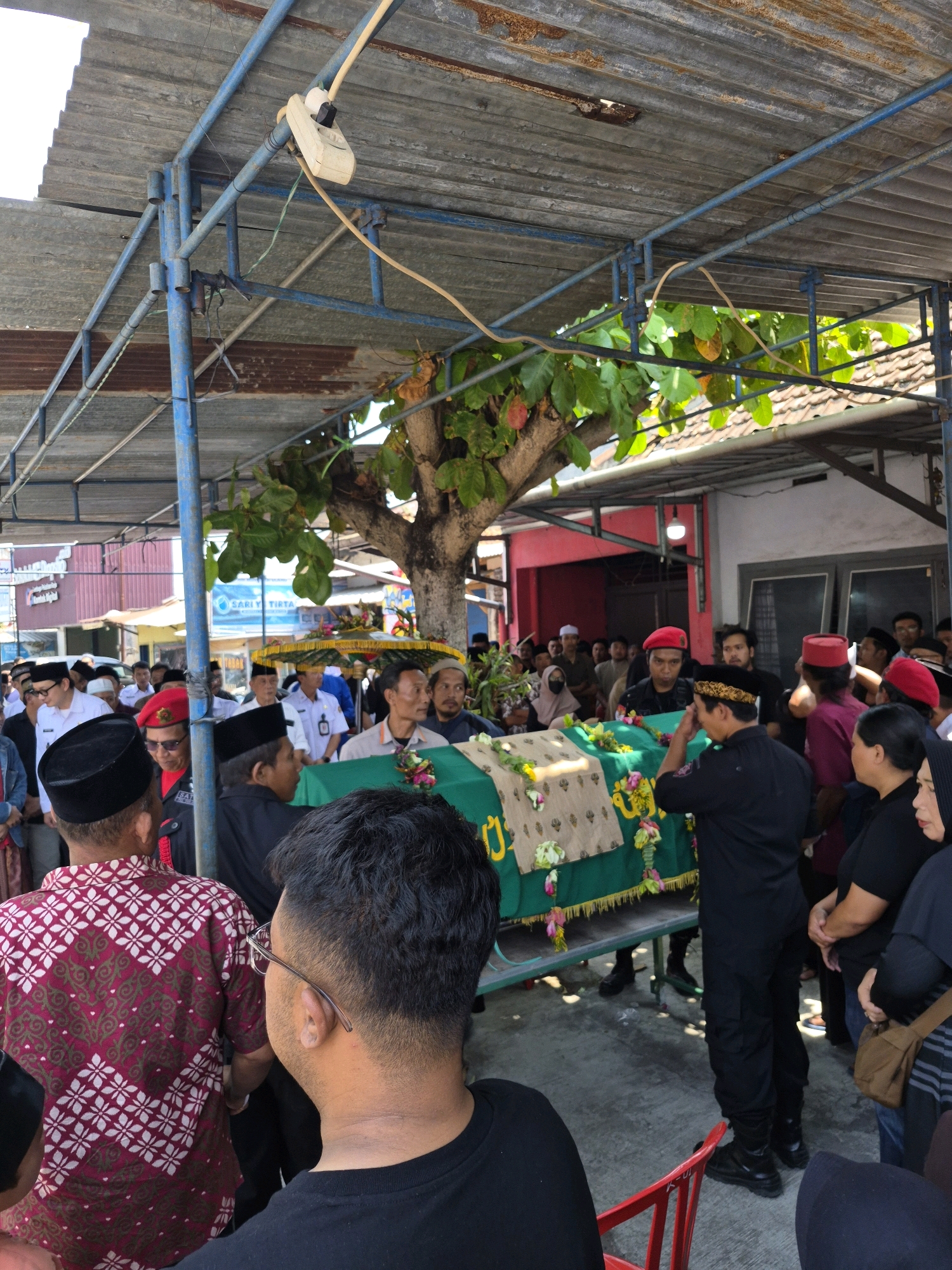 An outdoor funeral procession is depicted in Semawung Daleman, Indonesia, under a corrugated metal roof supported by blue metal poles. In the foreground, a man wearing glasses and a black t-shirt is seen from behind, his back towards the viewer. To his left, another man with dark hair is partially visible. Central to the image is a coffin draped in a green cloth, adorned with floral decorations. Several individuals surround the coffin, some wearing red hats, and others dressed in black attire, suggesting a somber and formal occasion. In the background, more people are gathered, and buildings with red and white facades are visible. The overall environment appears to be a community gathering space, possibly a cemetery or a community hall, given the presence of a makeshift shelter.