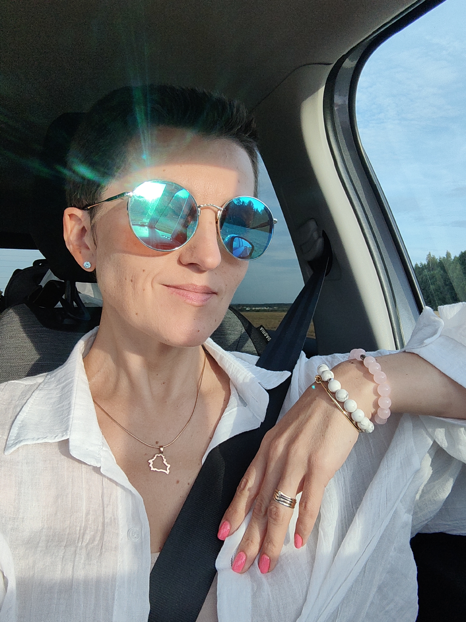 A woman with short dark hair and sunglasses is in the driver's seat of a car, wearing a white collared shirt and a delicate gold necklace with a map pendant. She is also wearing a bracelet with white and pink beads and a gold ring on her left hand. Her nails are painted bright pink. The car is on a road, with a blurry landscape of trees and fields visible through the window. The sky is blue with some clouds. The image appears to have been taken during the day. This is likely taken near Ilyanskiy selskiy Sovet, Belarus.
