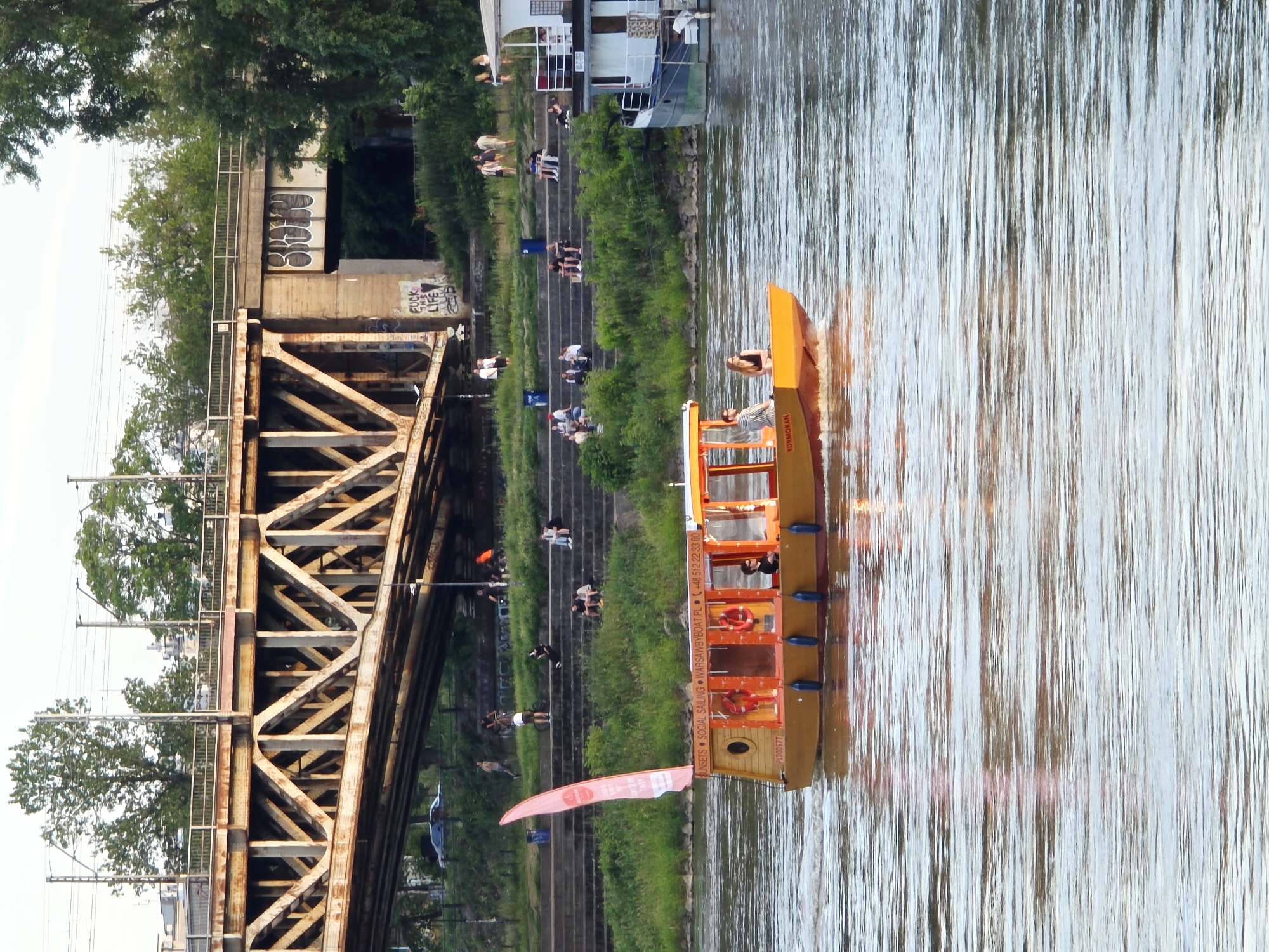 The image depicts a scene on the Vistula River in Warsaw, Poland, near the Poniatowski Bridge. A bright yellow tour boat, identified by the text "SUNSETS SOCIAL SAILING WARSAWBYBOAT.PL +48 512 22 33 00" on its side and "PL00057T" on its hull, is moving through the water. The boat has a covered cabin with large windows and an open seating area at the rear. Two people are visible inside the cabin, one driving and another seated. A pink flag with branding is attached to the top of the boat. In the background, the multi-level structure of the Poniatowski Bridge is prominent, with trains visible on the upper tracks. The riverbank is lined with grassy slopes and concrete steps, where several groups of people are seated and standing, enjoying the outdoors. Further to the right, another larger, white boat is docked. The sky is overcast, suggesting late afternoon or early evening light.