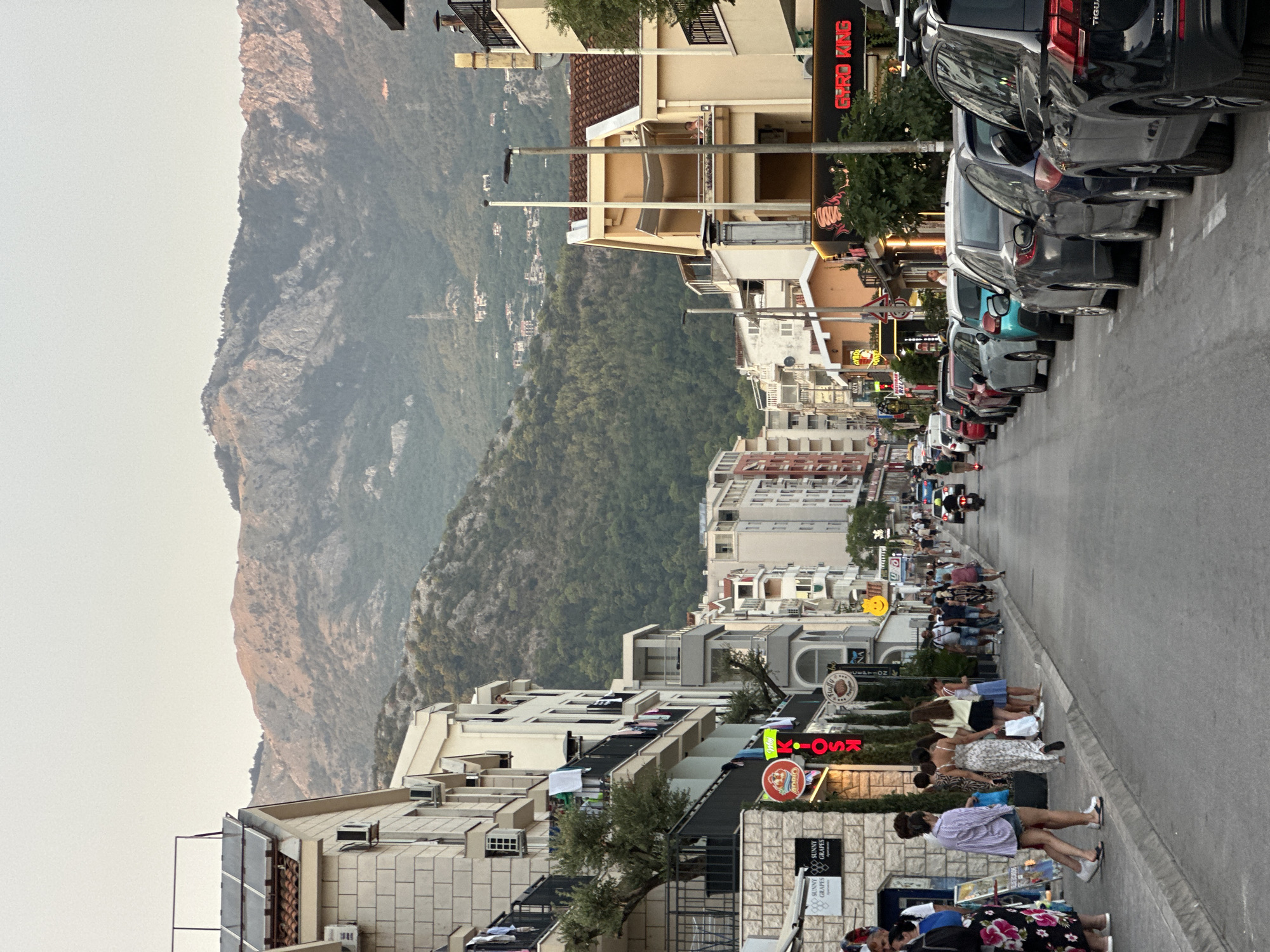 A street scene in Budva, Montenegro, features a mountain backdrop with lush green slopes and scattered buildings. The street is lined with multi-story buildings on both sides, displaying a mix of architectural styles and colors. The buildings house shops, restaurants, and residences, indicated by signs and visible windows. In the foreground, a sidewalk is populated with pedestrians walking and standing. Parked cars line the street on the right side. A motorcycle and a scooter are also present on the road. The overall atmosphere suggests a bustling urban environment during daylight hours, likely late afternoon given the soft lighting and long shadows.