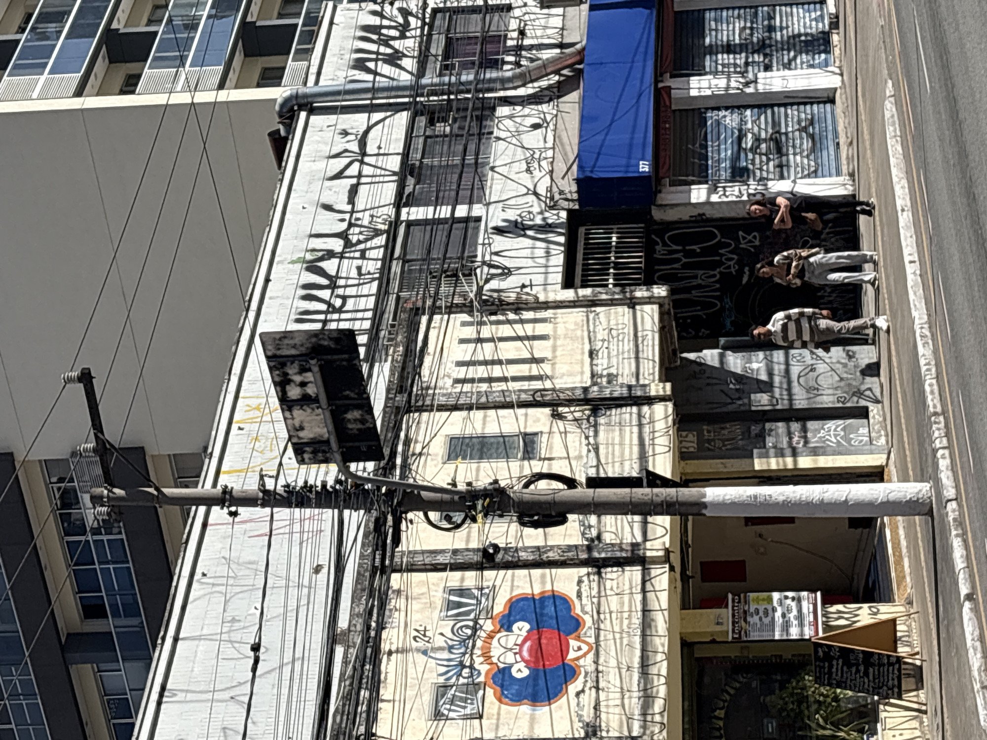 The image depicts a street scene in São Paulo, Brazil. A narrow street is visible with a sidewalk and road. On the left, a building with a clown face graffiti is present, with a menu displayed outside a store. To the right of this building is a utility pole covered in wires and electrical equipment. Further to the right, a larger building with multiple stories is covered in graffiti. Three individuals are walking on the sidewalk in front of the larger building. The building on the far right has several shutters. The overall scene is urban, with evidence of graffiti and a dense network of overhead electrical wires.