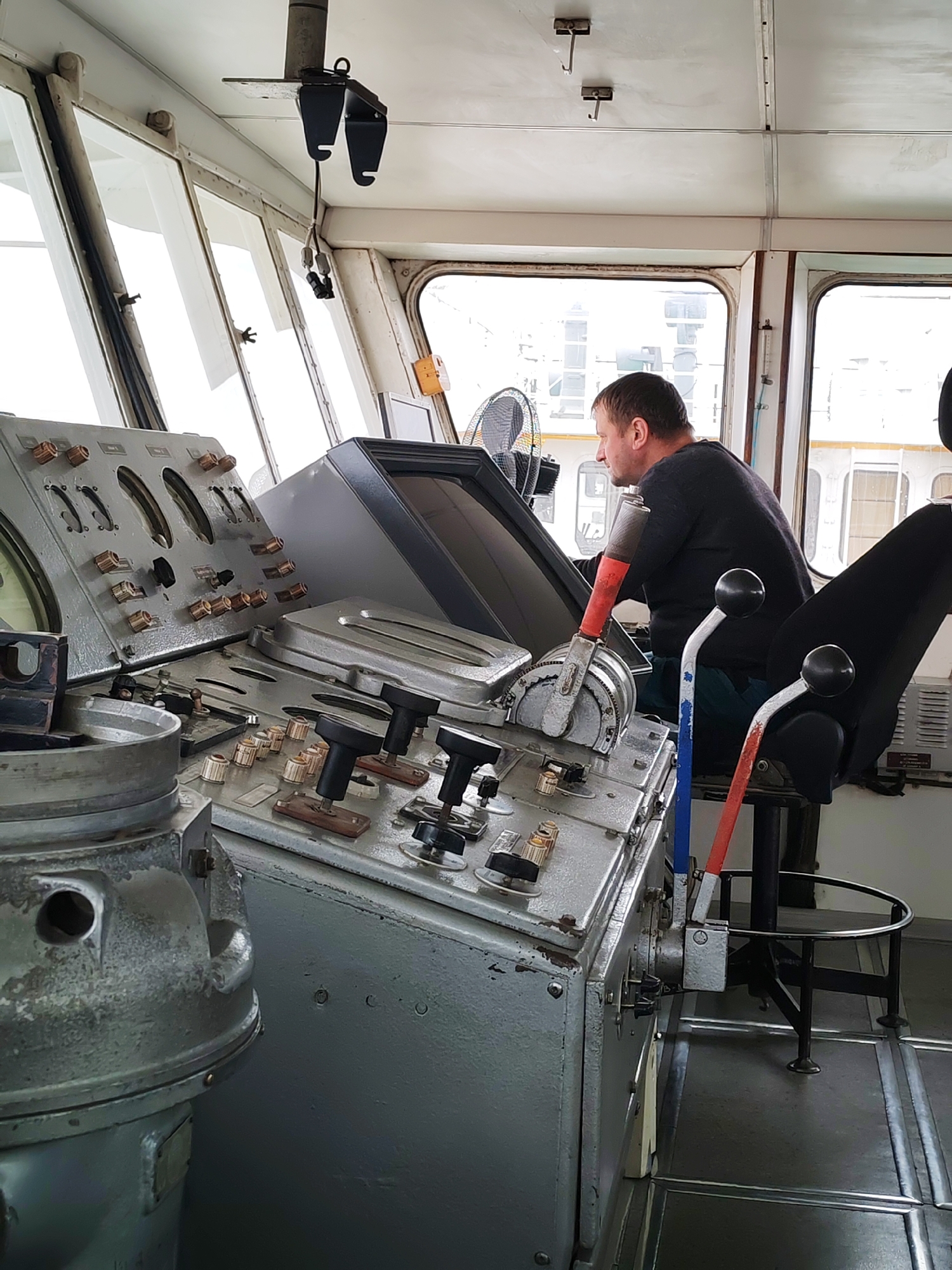 The image shows a man sitting in the control room of a ship or ferry, likely in Rybinsk, Russia. The man, dressed in a dark shirt and gloves, is operating the controls. The control panel is large and complex, with various knobs, levers, and gauges. The room is enclosed, with windows providing a view of the outside. The floor is metallic. The environment suggests an industrial setting.