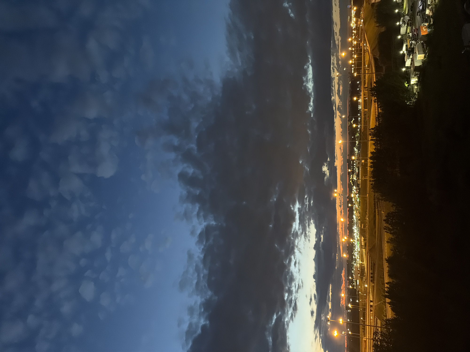 A wide shot shows an aerial view of a highway interchange and industrial area at dusk. The sky is a deep blue with scattered clouds that are dark and textured. The horizon is lit with the warm glow of streetlights and building lights from the city. The highway is visible with numerous lanes and overpasses, illuminated by tall lampposts. In the foreground, silhouetted trees obscure the view, and further down, a collection of parked trucks and trailers are visible, some with their lights on. The scene is devoid of people.
