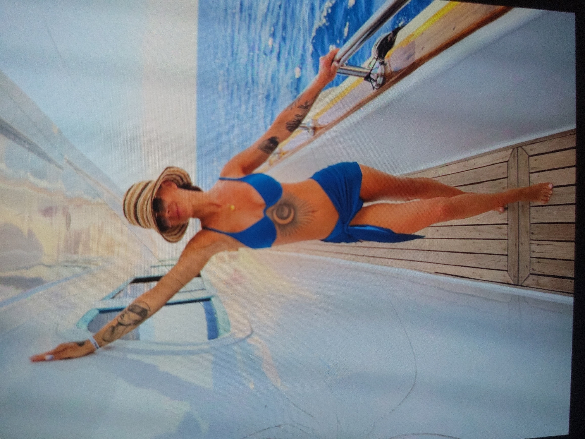 A vibrant outdoor shot captures a woman posing dynamically on a boat, likely a yacht, in Tsarevo, Bulgaria. She is positioned on her left side on the white hull of the vessel, extending her right arm upwards to grasp a metal railing and her left arm forward, resting her hand on the boat's surface. Her legs are stretched out to the right, resting on a wooden deck section.
The woman is attired in a bright blue two-piece bikini and a straw hat with striped detailing. She has visible tattoos, including a large, intricate sun-like design on her stomach and sleeve tattoos on both arms. A delicate golden necklace adorns her neck. Her fingernails are painted, and a light-colored bracelet is on her left wrist.
The scene takes place during a sunny day, with brilliant blue water visible in the background, suggesting open sea or a large body of water like the Black Sea. The boat features a sleek white exterior and a classic wooden deck. The image conveys a sense of confidence and relaxation, possibly from a vacation or a photoshoot. No discernible text is visible within the frame.