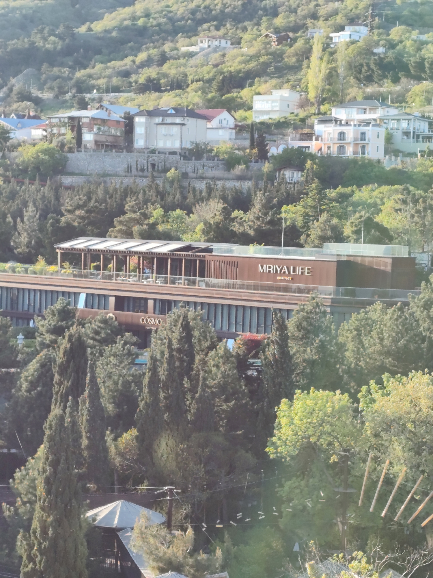 This medium-shot image captures a lush, green hillside densely populated with various buildings under natural light, likely during the day. The scene is set in Симеизский поселковый совет, Ukraine, showcasing a blend of natural landscape and developed structures.
In the mid-ground, a prominent modern building stands out with a multi-story, horizontal design. Its upper section features a rooftop terrace with a slatted wooden canopy, where small tables and chairs are visible, suggesting an outdoor dining or lounge area. Several figures appear to be present on this terrace. On the façade of this building, large golden letters clearly spell out "MRIYA LIFE INSTITUTE". Below this, on a lower floor of the same building, another sign reads "COSMO RESTAURANT". The building's architecture is sleek, with many vertical window panels.
Surrounding this main structure, the hillside is blanketed with numerous trees, including tall, slender conifers and broader deciduous trees. Scattered across the upper slopes are several residential-style buildings, varying in architectural design from modern white structures with flat roofs to more traditional homes with pitched, often tiled, roofs. These buildings are nestled among the greenery, some appearing to be perched on terraces or retaining walls built into the hill.
Towards the lower right, partially obscured by trees, elements of what appears to be a treetop adventure course are visible, with wooden planks and ropes strung between trees. In the bottom left, a gray, umbrella-like structure, possibly a gazebo or a small shelter, can be seen amidst more dense foliage. The overall impression is one of a scenic, developed resort or residential area integrated into a natural, hilly environment. The light is soft, suggesting either early morning or late afternoon, with no harsh shadows.