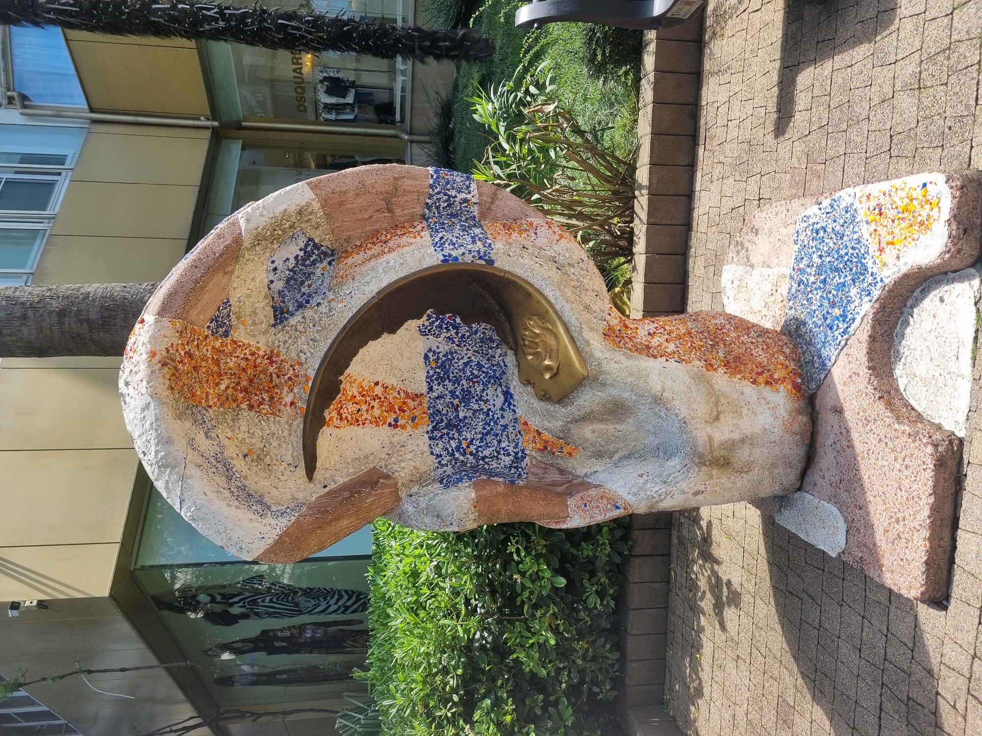 A stone sculpture of a human head with a bronze face is located on a paved walkway next to greenery, potentially in Sochi, Russia. The sculpture is made of rough stone with mosaic tiles in brown, white, orange, and blue patterns. A palm tree is visible behind the sculpture. A building with a glass facade is also behind the sculpture, with the word "DSQUARED" visible.