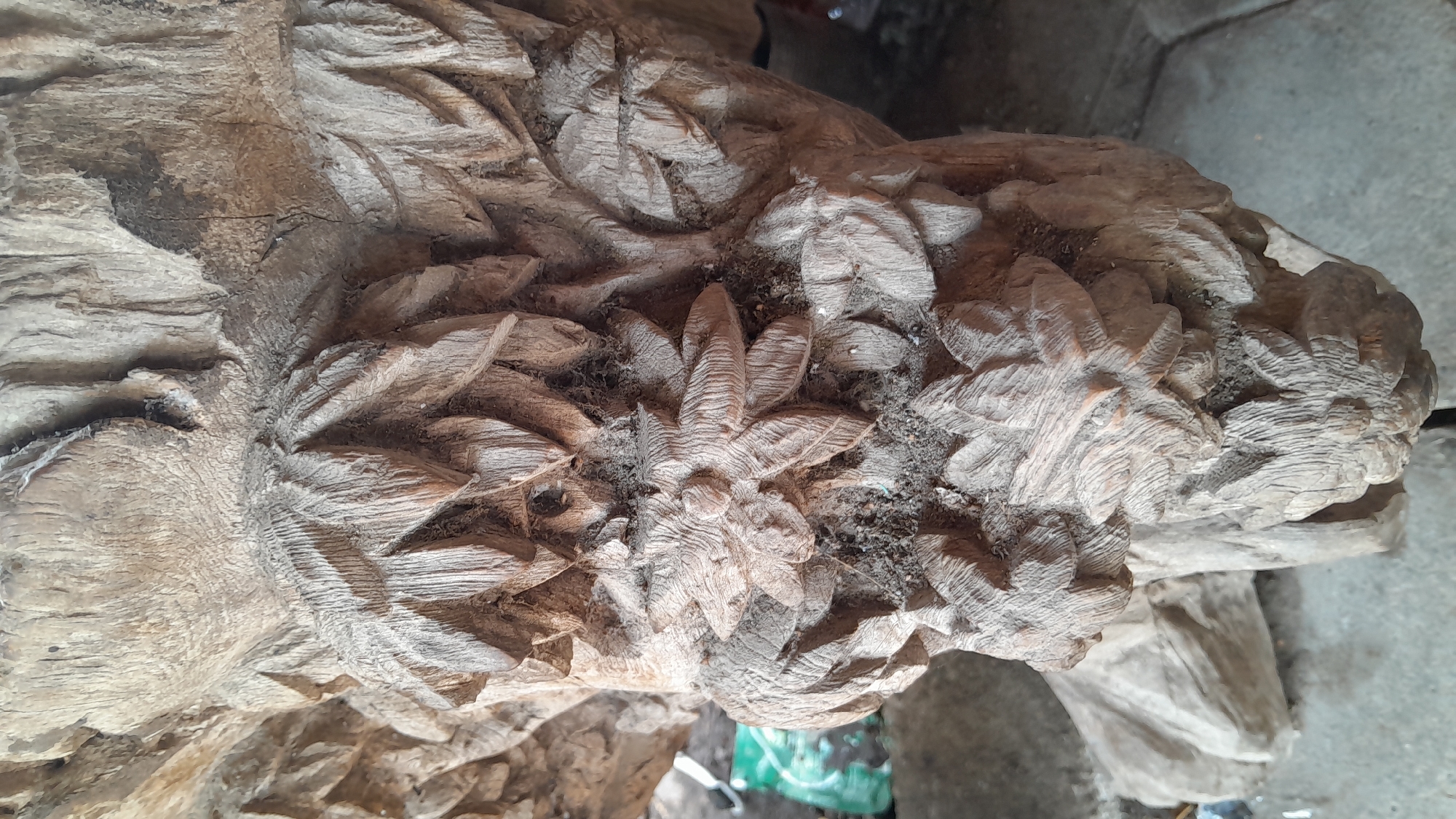 The image is a close-up shot of a carved wooden object, possibly a statue or furniture component, with intricate floral and leaf motifs. The wood appears aged and rough, with visible grain and some dust or dirt accumulated in the crevices of the carving. The lighting is somewhat diffused, casting soft shadows that highlight the texture of the wood. In the background, hints of a blurred environment are visible, including what appears to be a concrete floor and some green and dark colored objects, though their details are indistinct. The image does not contain any people or obvious actions, focusing solely on the carved wooden artifact. There is no information within the image itself to indicate its specific location in Sumberarum, Indonesia.