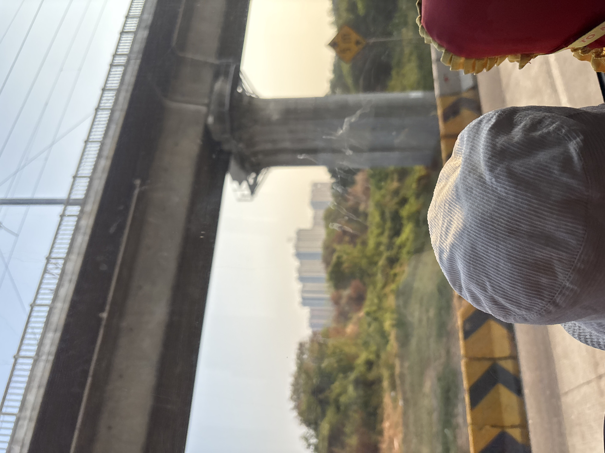 The image depicts a view from within a vehicle, looking outwards through a window. In the foreground, the rounded top of a light-colored, corduroy bucket hat is visible, along with a portion of a red object with decorative yellow trim on the right.
The middle ground features a road with yellow and black chevron barriers. Beyond the barriers, a dense area of green foliage and trees is present, with several tall, light-colored apartment buildings visible in the distance, suggesting an urban or semi-urban environment.
Dominating the upper half of the image is a large, elevated concrete structure, likely a bridge or overpass, supported by massive concrete pillars. Power lines or cables are visible crisscrossing the sky above the structure. The sky is a pale blue with hints of yellow, indicating either early morning or late afternoon light. A yellow diamond-shaped sign with a black arrow indicating a curve is partially visible on the right side of the image, near the concrete pillar.
The location is identified as Jayamukti, Indonesia. The scene suggests a journey, possibly on a highway or main road, passing through a developing area where urban infrastructure, like elevated transport routes and residential buildings, is present alongside natural green spaces.