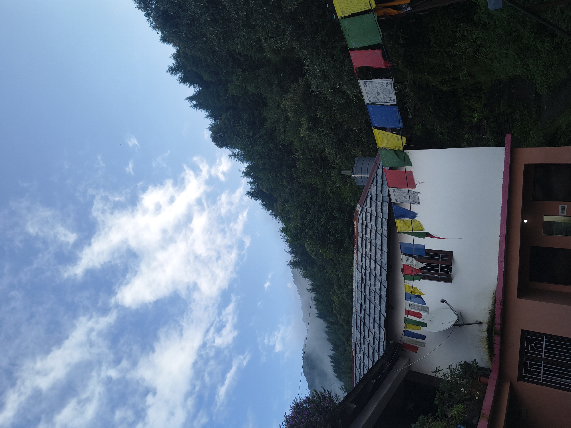 In Haini, India, a whitewashed building with a slate roof is adorned with a string of colorful prayer flags that stretch across its facade. The flags, bearing Tibetan script, are a vibrant mix of blue, white, red, green, and yellow, fluttering gently in the breeze. A satellite dish is mounted on the wall below the flags, and a dark-tiled roof extends from the upper portion of the building. The building is nestled against a densely forested hillside, with tall, dark green trees creating a dramatic backdrop. In the distance, misty mountains rise, partially shrouded in clouds, against a clear blue sky dotted with wispy white clouds. The lower section of the building is painted a soft coral color and features dark-framed windows.