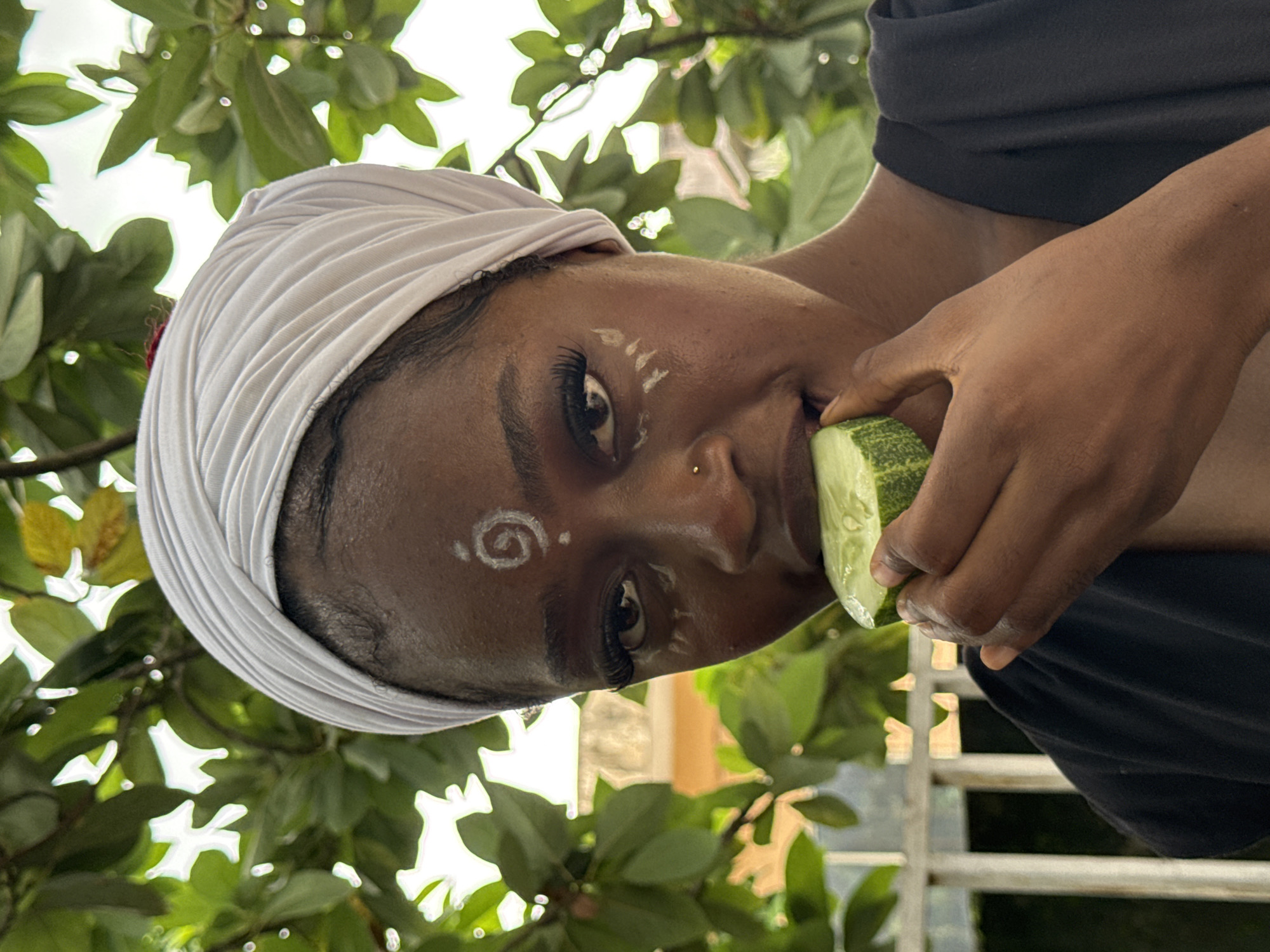 In Ado, Nigeria, a Black woman with a white headscarf is captured in a close-up, medium shot, eating a slice of cucumber. Her face is adorned with white face paint, including a spiral on her forehead and markings below her eyes. She wears a black garment. The background is a blur of lush green foliage, suggesting an outdoor setting amidst nature.