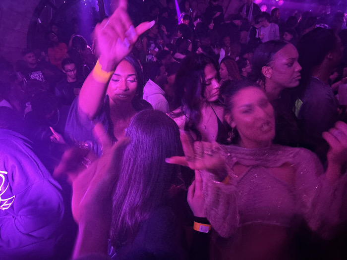 A crowd of people is gathered in a dimly lit space with vibrant purple lighting. Many are dancing and moving their hands. A woman in a dark top with a yellow wristband raises her hands. To her right, another woman in a sparkly top is also dancing. In the background, multiple faces are visible amidst the crowd, suggesting a lively and social atmosphere.