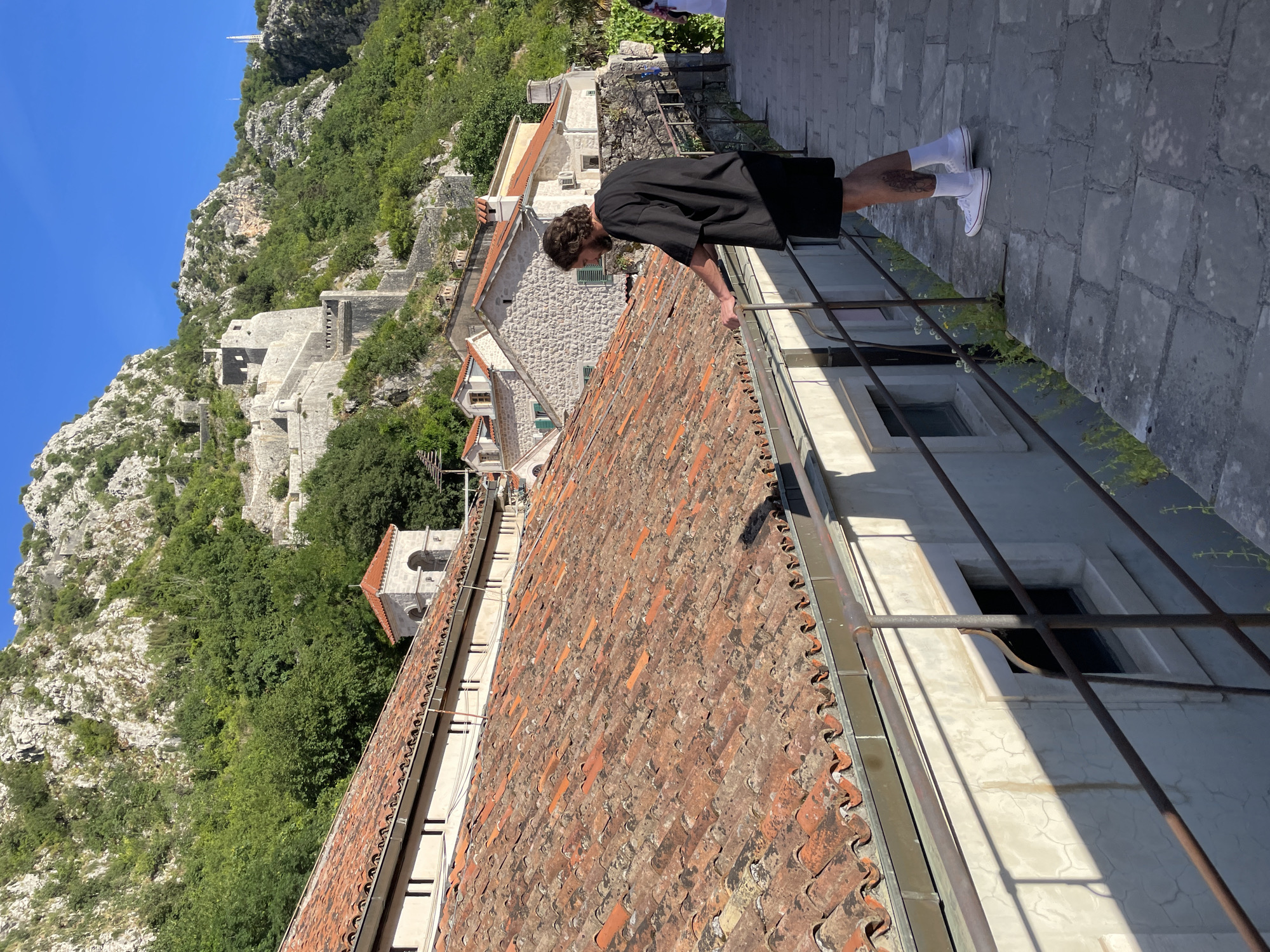 In Kotor, Montenegro, a man with curly hair, wearing a black t-shirt, black shorts, white socks, and white sneakers, stands on a stone pathway, leaning on a metal railing. He is looking down and to his left. Behind him, the pathway curves to the right, bordered by a low wall with windows and an aged, red-tiled roof. In the background, stone buildings with terracotta roofs cluster together, and beyond them, a steep, green mountain rises, crowned by ancient stone fortifications. The sky above is a clear, bright blue.