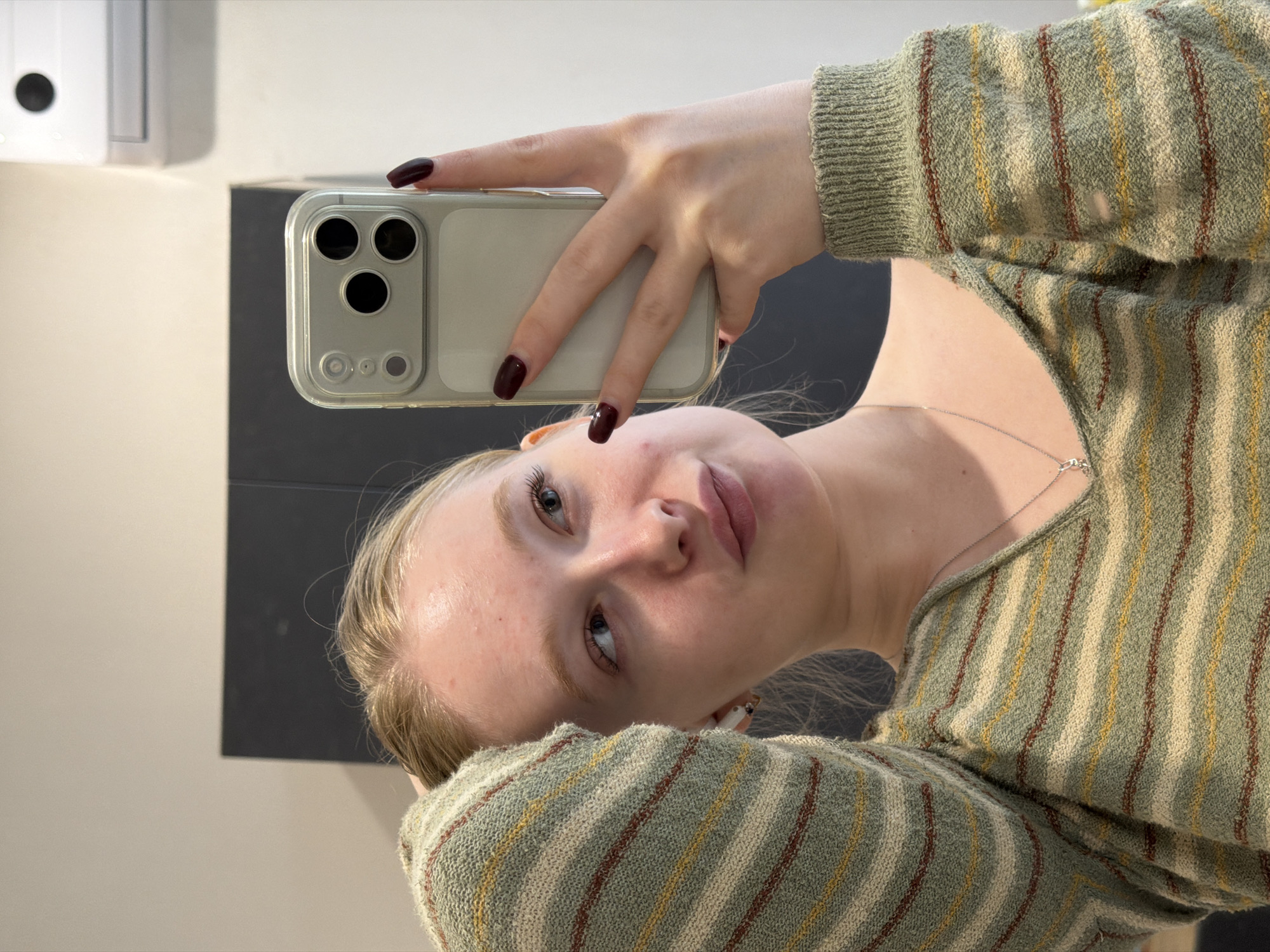 A young person with fair skin and blonde hair is holding a phone with three rear cameras. The person is wearing a striped, green and yellow sweater and has dark nail polish. A silver necklace is visible around the neck. The background includes a wall and a dark, rectangular shape. No specific location in Засечное, Russia is identifiable based on the image.