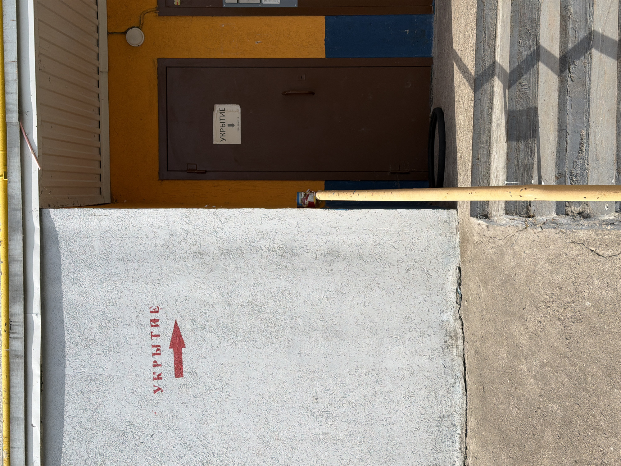 The image shows an exterior scene in Naberezhnye Chelny, Russia. An arrow and the word "УКРЫТИЕ" (shelter) are painted in red on a white concrete wall, indicating the location of a shelter. A brown metal door, possibly the entrance to the shelter, is set into a yellow and blue wall above the concrete wall. A yellow pipe runs horizontally across the frame. Stairs are visible to the right of the door. The scene appears to be in daylight, with shadows cast by the stairs. The environment is urban and utilitarian.