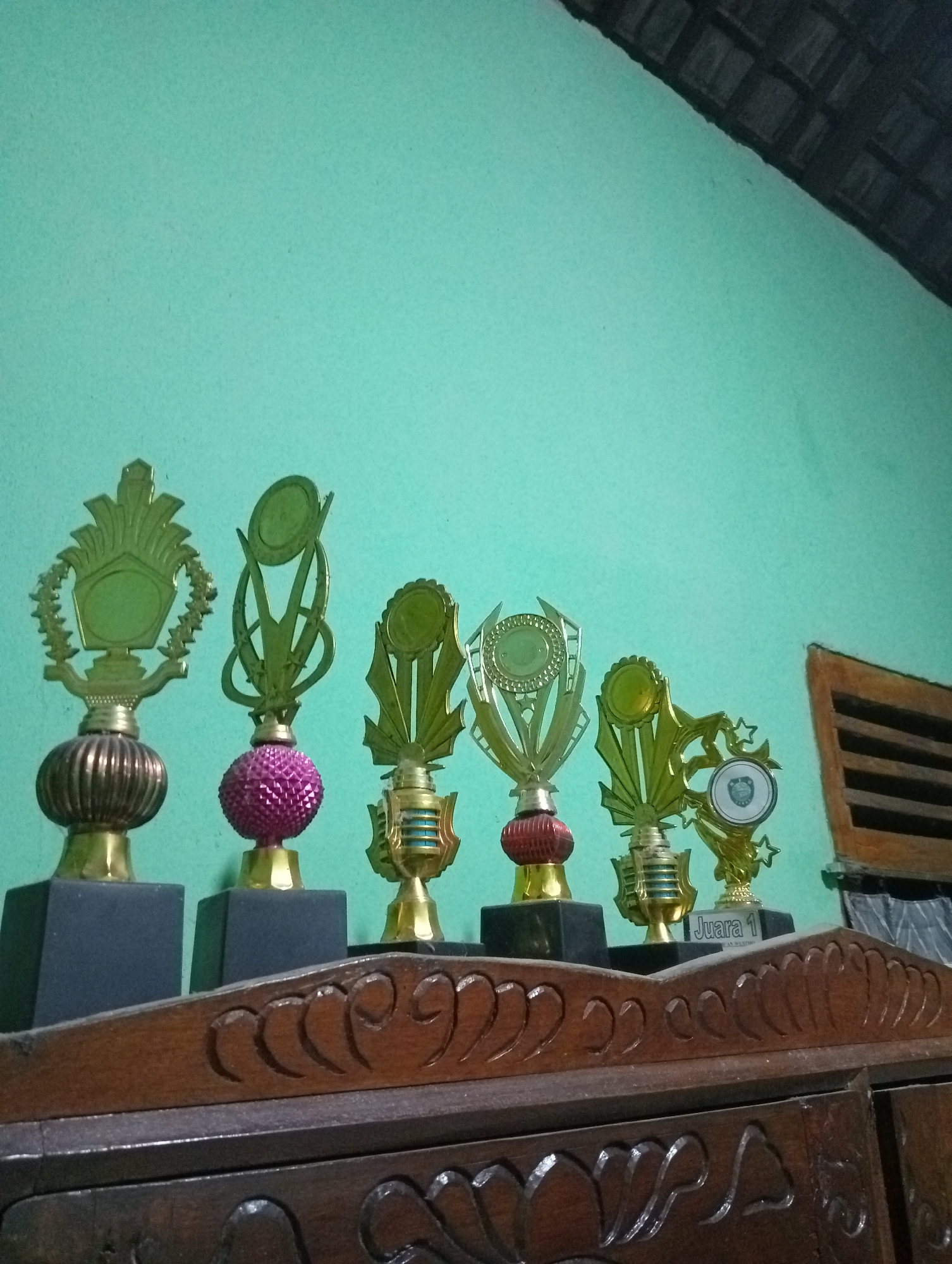 A collection of six gold trophies of varying sizes and designs are displayed on a dark wooden shelf against a light teal wall. The trophies are topped with decorative elements and some have colored accents. From left to right, the first trophy has a ribbed gold base and a flat, ornate gold topper with a circular indentation. The second trophy has a dark, ribbed base and a vibrant pink, spherical element below a gold topper. The third trophy is smaller, with a gold base and a topper featuring green and blue striped details. The fourth trophy has a red, ribbed base and a gold topper with a circular emblem. The fifth trophy is similar to the fourth but with a gold topper that has star embellishments and a circular emblem. The sixth trophy is the smallest and has a gold base and a gold topper with a circular emblem. Below the trophies, the shelf has carved decorative details. To the right of the trophies, a wooden grate or blind is visible against the teal wall. The scene appears to be indoors, likely in a home, with the lighting suggesting daytime. The trophies imply a setting where achievements or competitions are recognized, possibly in a school or community context in Kota Negara Timur, Indonesia. One trophy clearly displays the text "JUARA 1", meaning "Champion 1" in Indonesian.