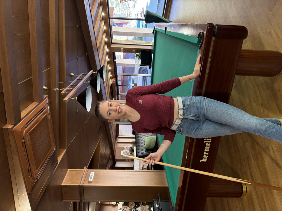A young adult female stands in an indoor lounge or game room located in Kladnitsa, Bulgaria. She is positioned to the left of a dark brown wooden pool table with a green felt surface, upon which "hermeli" is inscribed. The woman, with dark hair pulled back and a light complexion, wears a maroon cable-knit V-neck crop top, light blue denim jeans, and a white belt with a gold "G" buckle. She holds a light-colored wooden pool cue vertically in her left hand, while her right hand rests on the edge of the pool table. She has dark nail polish and wears a white bracelet on her left wrist and a red and white bracelet on her right. Her expression is a direct smile towards the viewer.
The room features a wooden parquet floor and a dark wooden ceiling with exposed beams. A square, recessed air conditioning unit is visible in the ceiling. Above the pool table, a fixture with three black cone-shaped pendant lights hangs. In the background, to the left, a dark green upholstered booth is visible, along with a foosball table and an "EXIT" sign on a wooden pillar. To the right, large windows reveal an outdoor scene with parked vehicles and what appears to be a sunny day. A dark armchair is also visible near the windows.