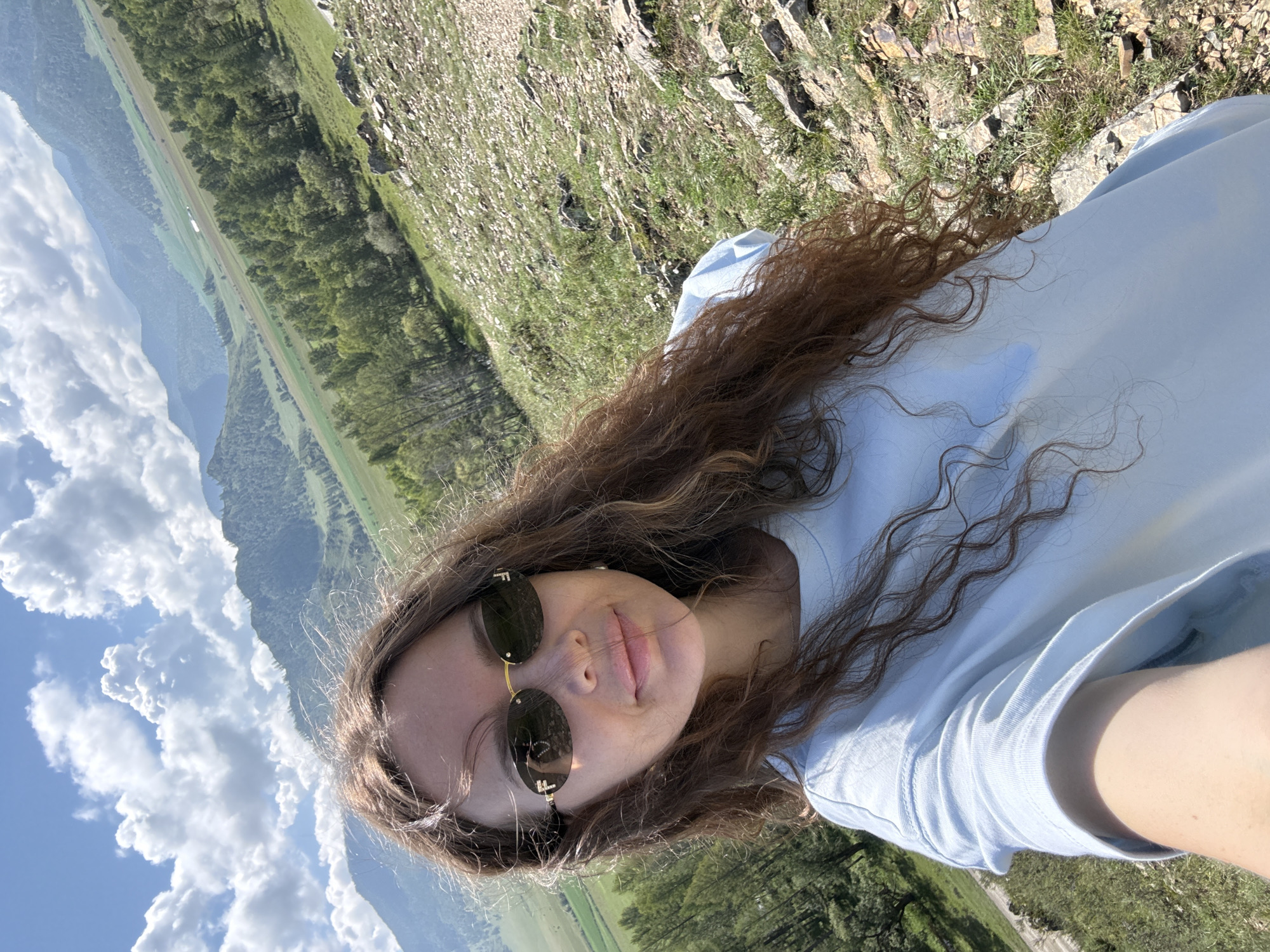 A young woman with long, wavy brown hair and sunglasses is taking a selfie outdoors. She is wearing a light blue t-shirt. The background shows a mountainous, green landscape under a blue sky with white clouds. The terrain in the foreground is rocky and covered with grass. The location appears to be Bichiktu-Boom, Russia.