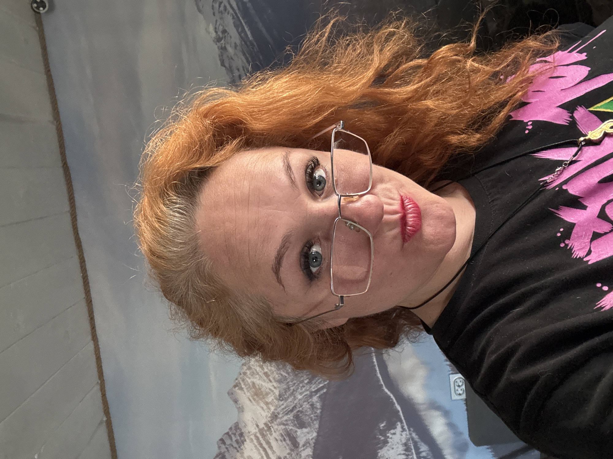 A woman with red hair and wearing glasses is positioned in front of a mountain landscape backdrop. She is wearing a black t-shirt with pink designs. A necklace with a gold-colored pendant is visible around her neck. The backdrop features a mountain range, presumably in Chaikovsky, Russia. The lighting appears to be indoor and diffused.