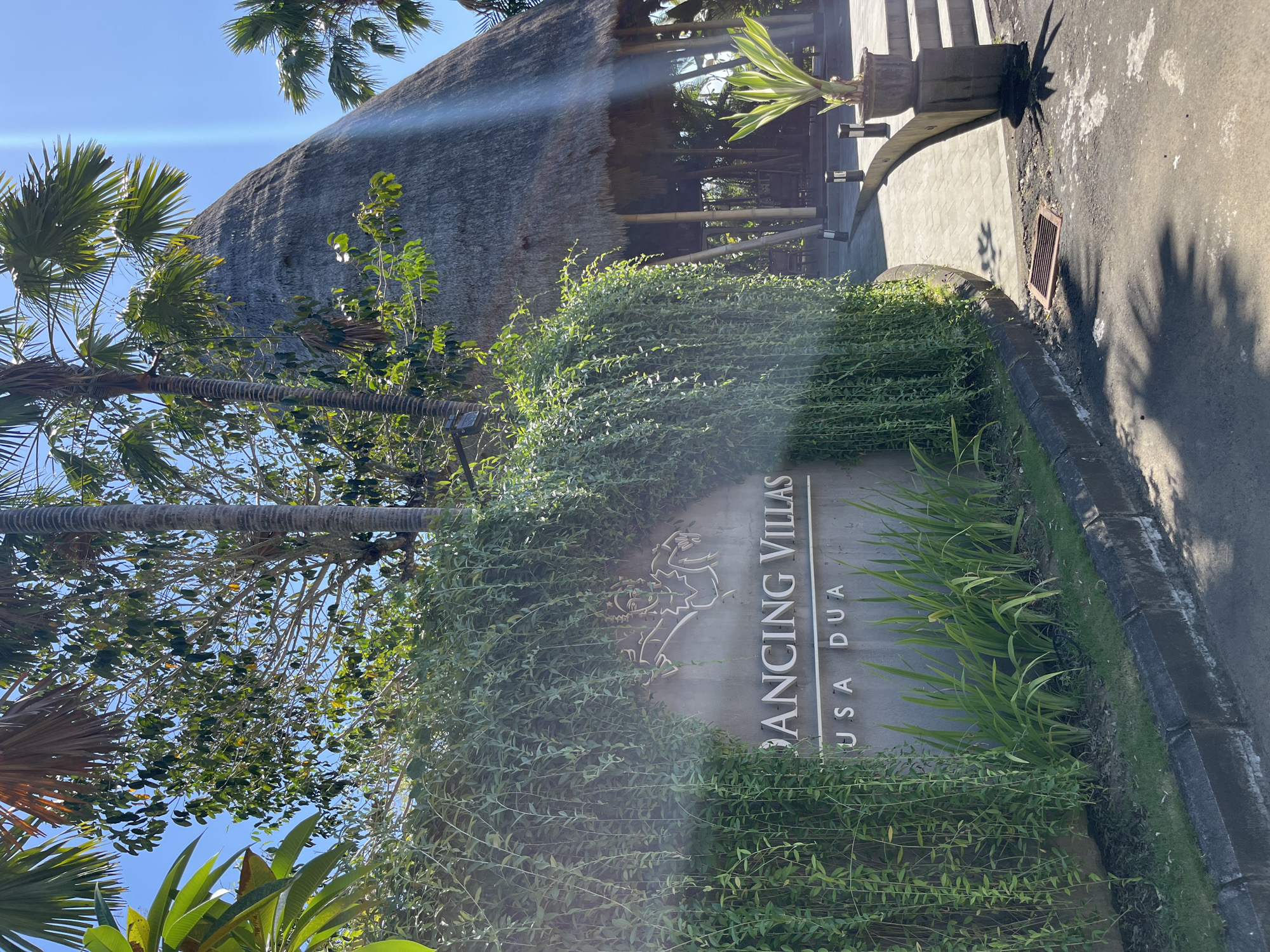 In Kuta Selatan, Indonesia, a sign for "DANCING VILLAS NUSA DUA" is prominently displayed, partially obscured by hanging green vines. The sign is made of grey stone with the text and a stylized depiction of a dancer etched into it. Behind the sign, a large thatched-roof building with bamboo supports is visible, along with palm trees and lush greenery. The scene is bathed in sunlight, casting shadows on the paved walkway and road in the foreground. The environment appears to be a resort or a similar hospitality establishment.