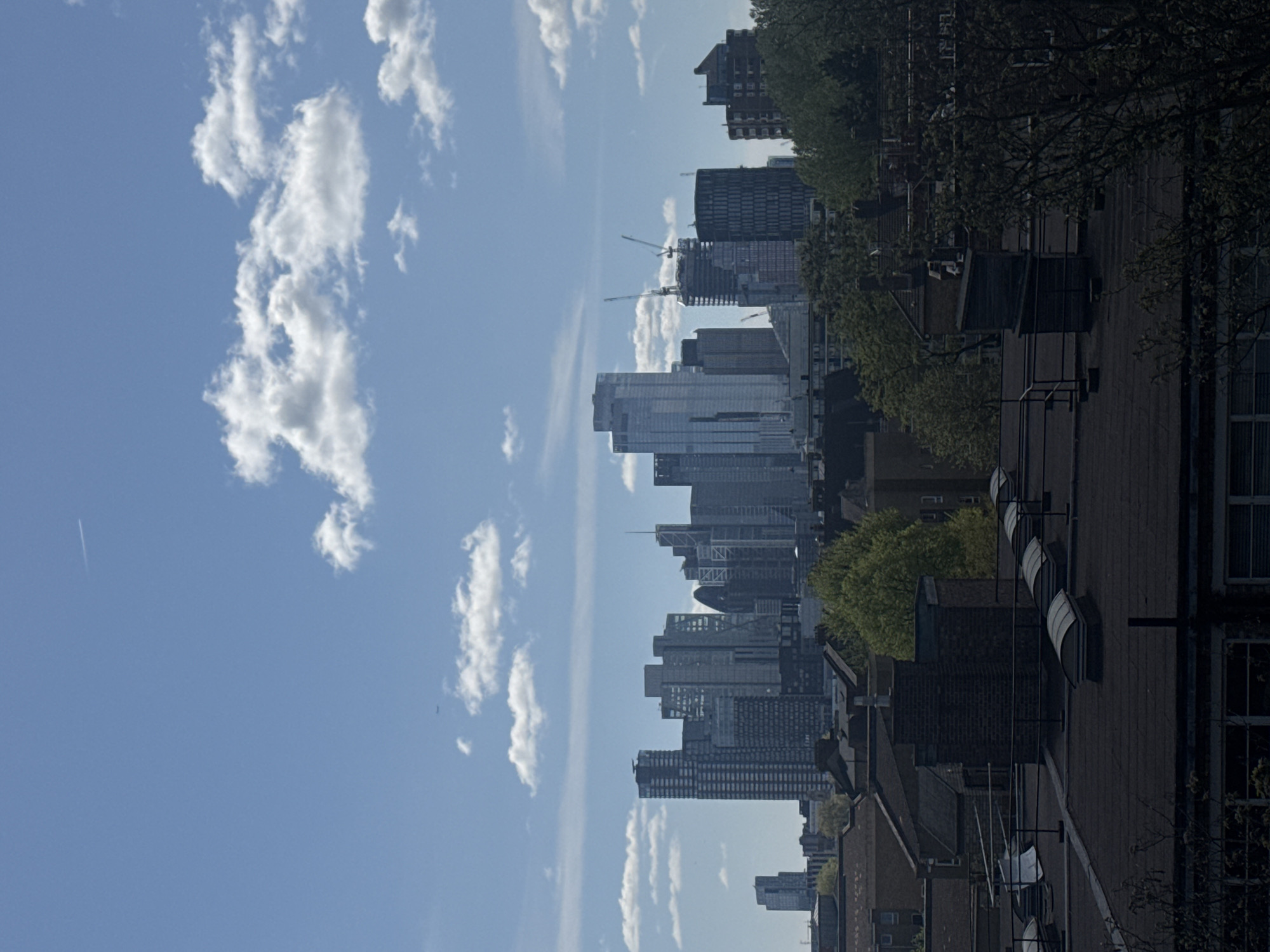 The image captures a skyline view in London, United Kingdom, juxtaposing residential rooftops with modern skyscrapers. The sky is a bright blue with scattered white clouds. Tall buildings, including several under construction with visible cranes, dominate the cityscape. Adjacent to the cityscape, there are rooftops of residential houses and trees with early spring foliage.