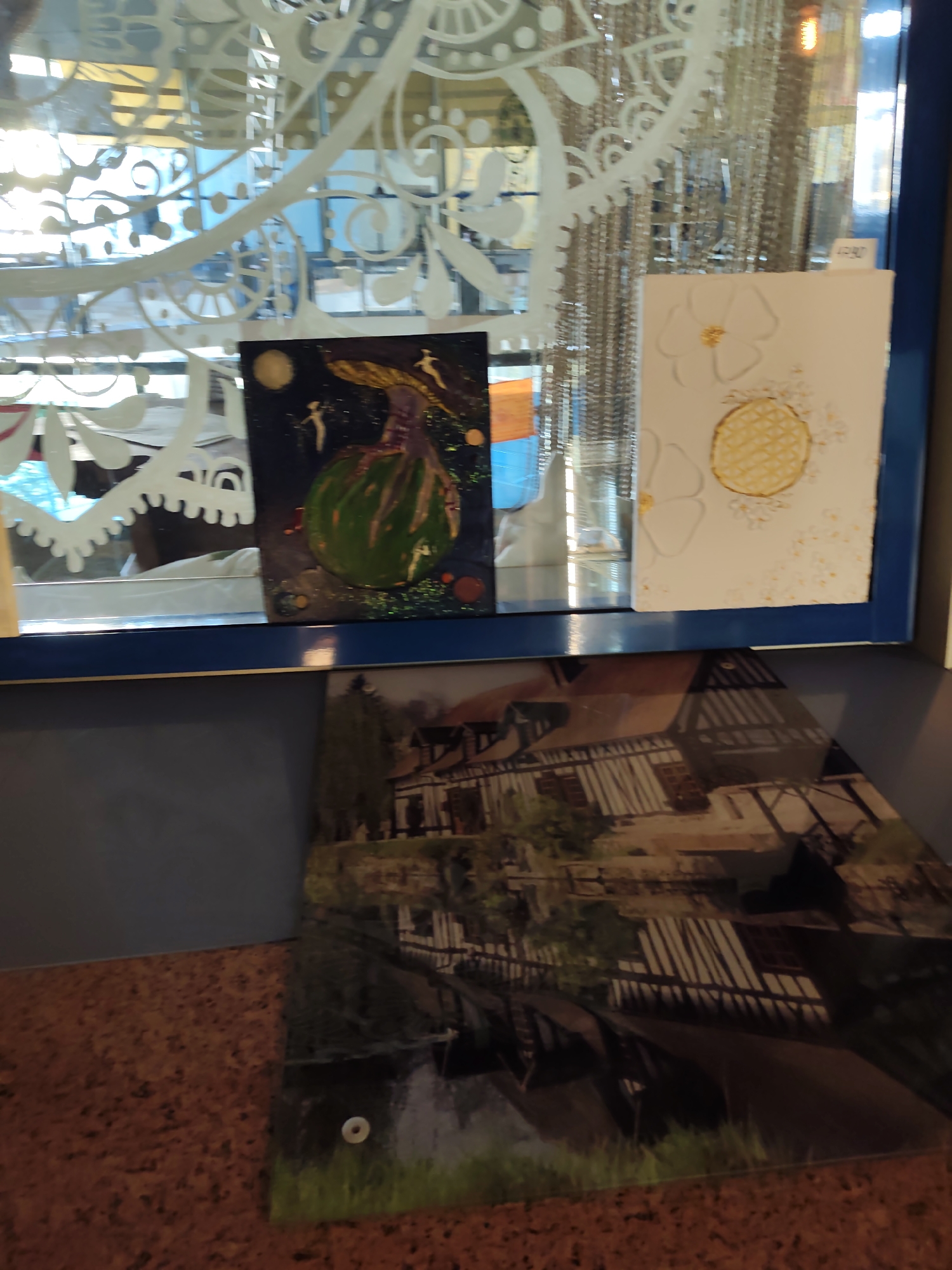 In the foreground, a cork-textured surface holds a large, glossy print featuring a reflection of a building with a dark roof and arched windows, partially obscured by shadows. The print is attached with a small white circular fastener. Above this, on a blue shelf, sit two small framed artworks. The one on the left is a dark square painting depicting a green, bulbous object under a yellow moon, with small white figures scattered around it. The artwork on the right is a white rectangle with a large white flower and a circular, textured yellow design. Behind the artworks, a decorative, white, ornate pattern is visible against a blurred background of light and what appear to be vertical strands. The overall scene suggests an indoor display of art. The image does not contain enough information to determine the location in the city of Sochi, Russia.