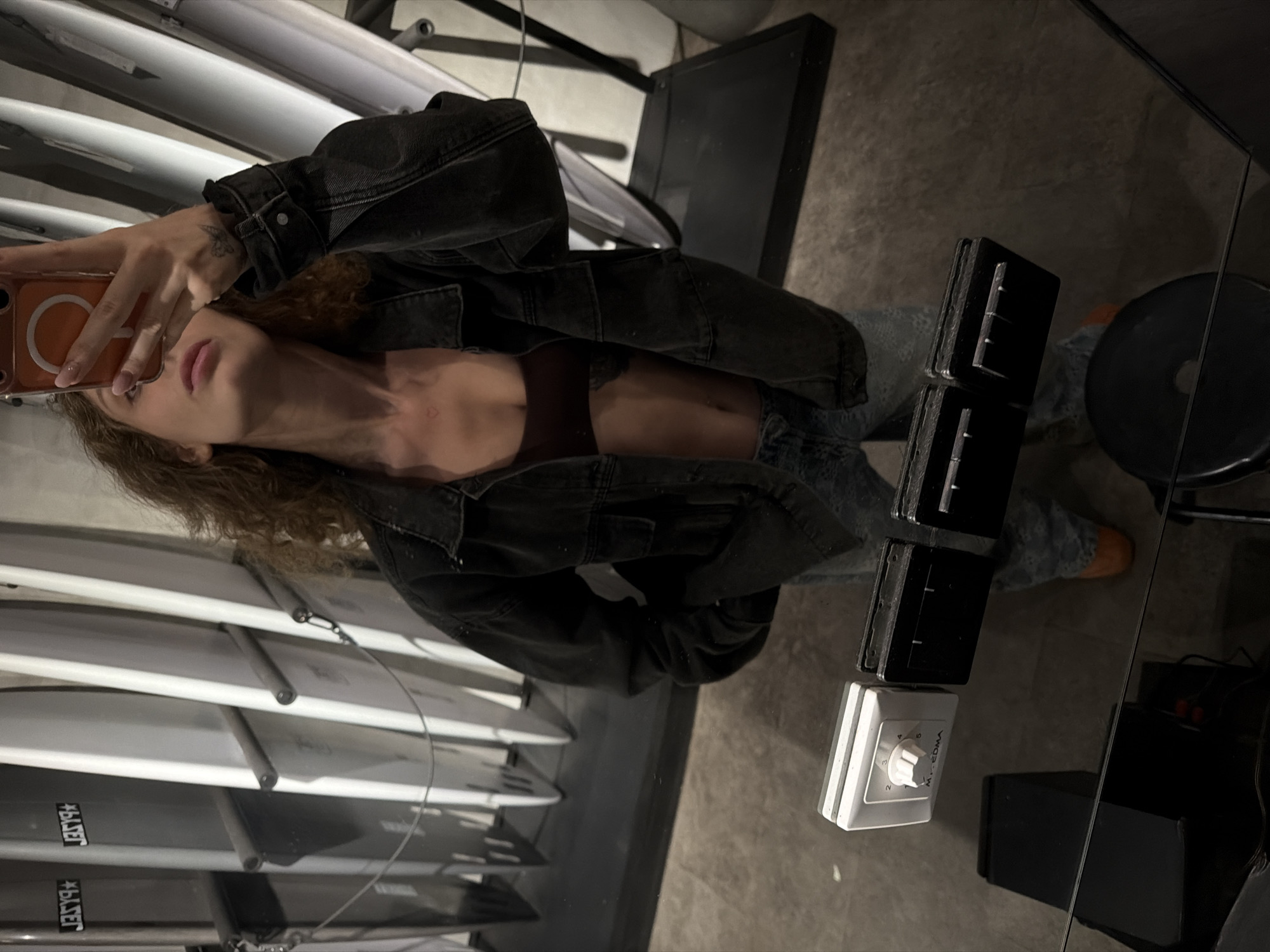 A person is taking a mirror selfie in what appears to be a retail store or showroom. The individual is wearing a dark denim jacket, a dark maroon bralette, and light-wash jeans. Their hair is curly and brown. In the background, there are several white surfboards arranged vertically. In the foreground, a dark reflective surface, likely a mirror, shows the person's reflection and a portion of the floor. Attached to the mirror are three black magnetic clips and one white switch with a dial labeled "Mr. EDMA". The lighting suggests an indoor environment, possibly evening. The scene is in North Kuta, Indonesia, a region known for its surf shops and proximity to beaches.
