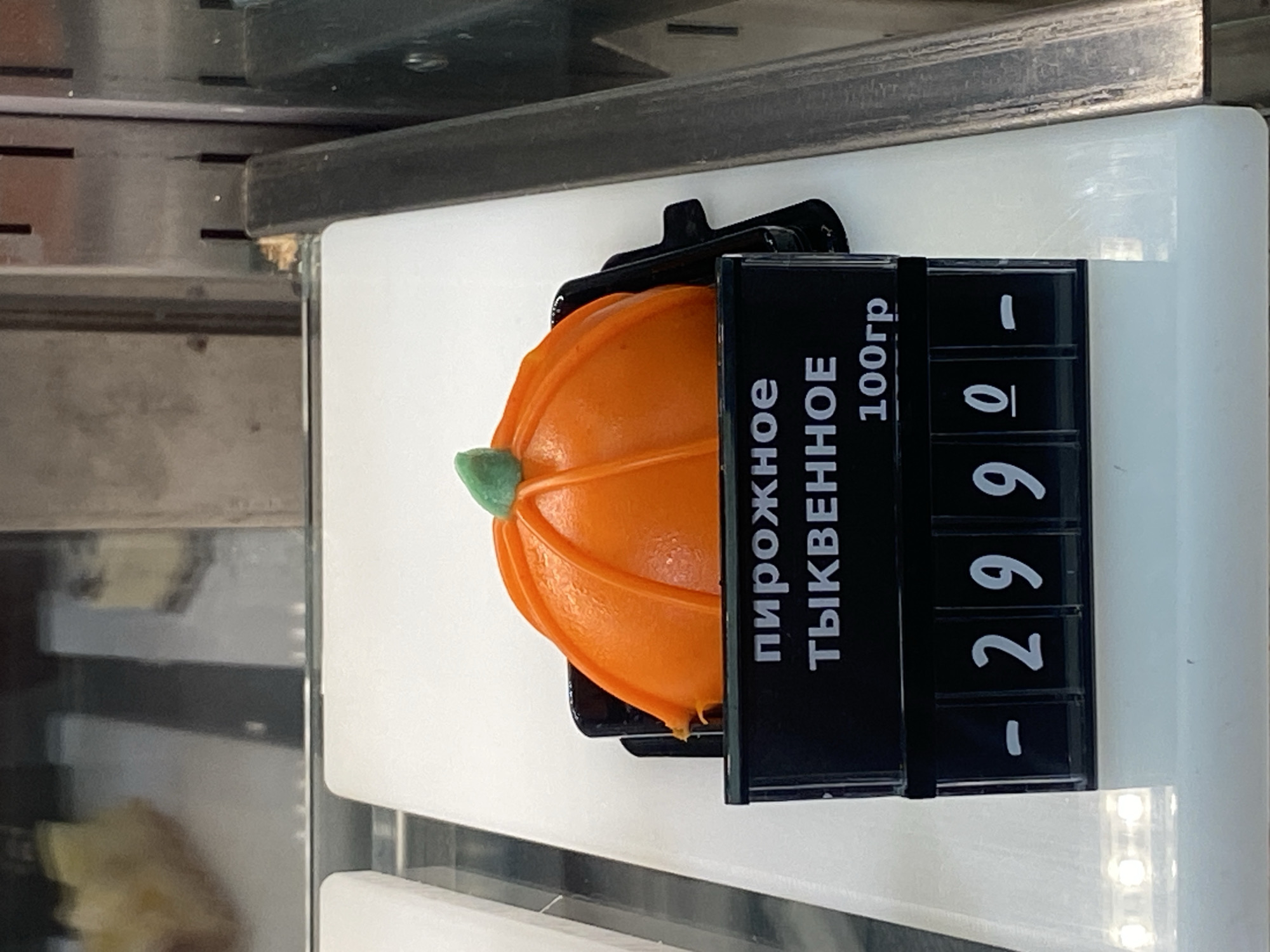 The image displays a single, vibrant orange pastry, meticulously shaped like a pumpkin, presented for sale in what appears to be a retail display case. The pastry features distinct vertical ridges, typical of a pumpkin, and is topped with a small, light green, stem-like embellishment. It rests within a dark, possibly black, individual serving container on a clean, white shelf or display surface.
Positioned immediately to the right of the pastry is a black price tag holder featuring white text and numbers. The text, written in Russian, clearly identifies the item as "ПИРОЖНОЕ ТЫКВЕННОЕ" (Pirozhnoe Tykvennoe), translating to "Pumpkin Pastry" or "Pumpkin Cake". Below this description, "100гр" (100gr) indicates a portion size of 100 grams. The price is displayed as "- 2990", which in the context of Russia (Sochi), signifies 299.0 Rubles.
The setting suggests a commercial environment such as a bakery, cafe, or grocery store, likely within a refrigerated unit, indicated by the metallic elements in the background. The pumpkin shape of the pastry strongly implies a seasonal offering, possibly for autumn or holidays like Halloween or Thanksgiving. No individuals are visible in the image.