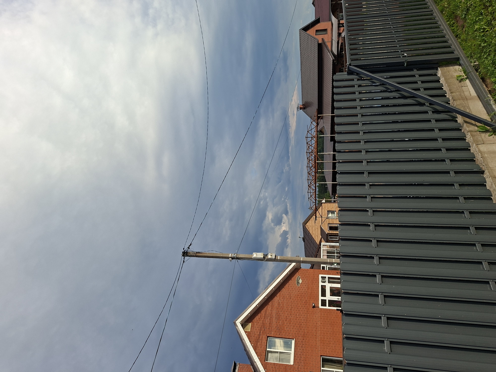 The image depicts an outdoor scene with a cloudy blue sky. In the foreground, a dark gray metal fence runs horizontally across the frame. Behind the fence, there are several houses with red brick and brown siding, and dark brown roofs. Power lines and a utility pole with electrical equipment are also visible, stretching across the sky. The scene appears to be in a residential area, possibly in a suburban or rural setting, given the presence of houses and the natural elements.
The lighting suggests it is daytime, with the clouds obscuring direct sunlight and creating a diffused light. The weather appears to be partly cloudy. There is no discernible activity or event taking place; it seems to be a static view of a landscape. No people are visible in the frame.
Notable details include the modern design of the houses with their distinct roofing and window styles. The metal fence is a prominent feature, suggesting a boundary or enclosure. The presence of power lines indicates an established infrastructure. The location context provided is Прикамский, Russia. There is no visible text in the image.