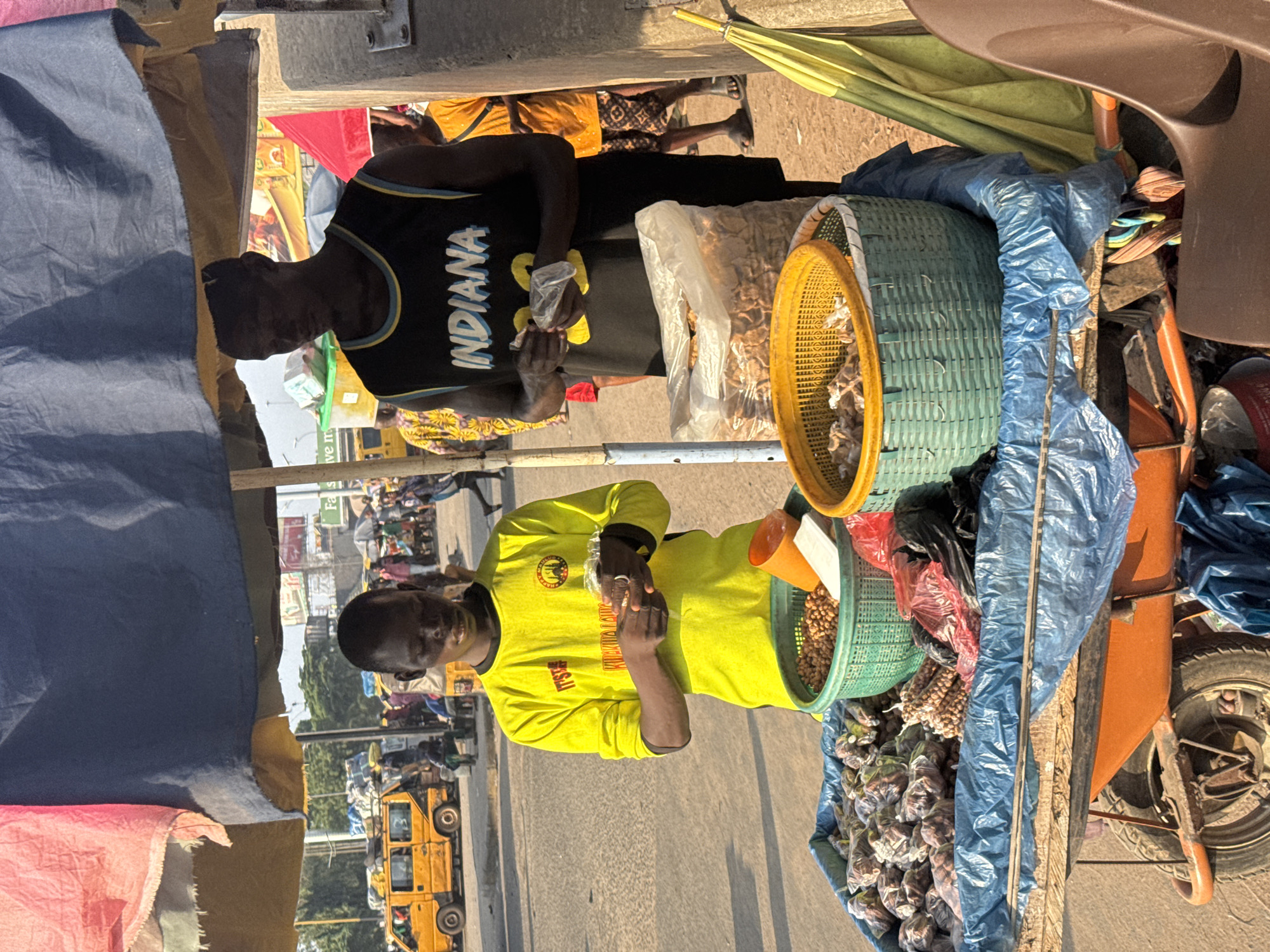 The image depicts a street scene in Onitsha, Nigeria, specifically at a roadside market. Two young men are visible. The man on the left wears a bright yellow long-sleeved shirt with inscriptions. He is focused on handling small items, possibly food products, within plastic packaging. The man on the right wears a black sleeveless t-shirt with "INDIANA" and the number "23" printed on it. He is also handling small items in plastic.
In front of them is a mobile cart laden with various food items, primarily nuts and other snacks, packaged in plastic bags and displayed in woven baskets. The cart is covered by a blue tarp, with additional awnings made of fabric in red and dark blue. Behind the men, the bustling activity of the city is evident, with a yellow minibus and other vehicles on the road, along with pedestrians and other market stalls. The ground appears to be paved, suggesting a main thoroughfare within the city. A green umbrella is visible to the right of the cart. The lighting suggests it is daytime, possibly late afternoon due to the long shadows.