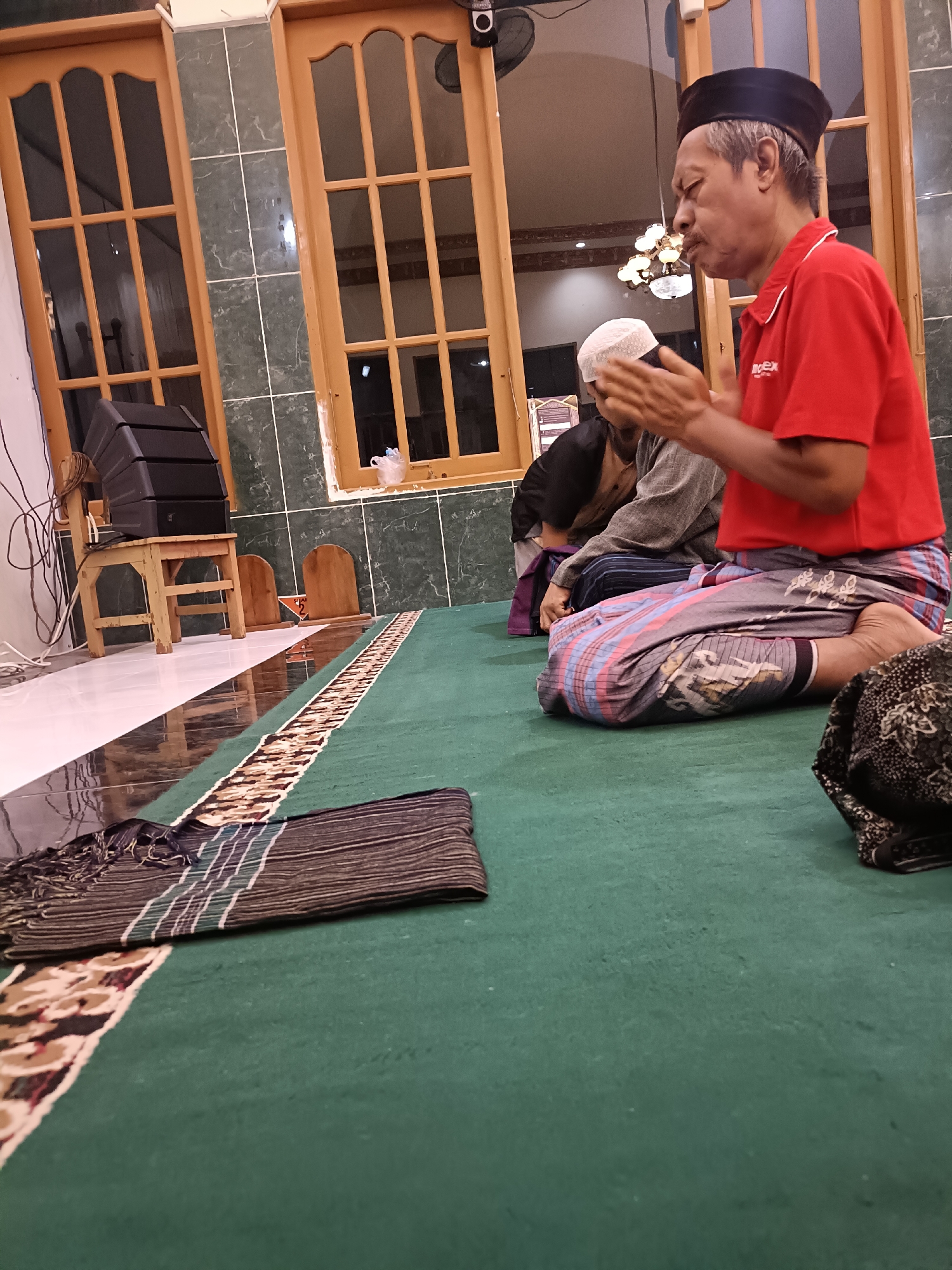 The image depicts a scene inside a mosque in Kediri City, Indonesia. Two men are visible, kneeling on a green carpeted floor. The man in the foreground, on the right, is wearing a red polo shirt with a logo and a black peci (cap). He has his hands clasped together at chest level, and his eyes are closed. He is wearing a sarong with a colorful striped pattern. The second man, partially visible behind him and to the left, is wearing a white peci and a grey shirt. He is also kneeling, facing downwards. The environment includes wooden window frames with glass panes, green tiled walls, and a patterned rug on the floor. A speaker is mounted on a wooden stool in the background, along with some wires.