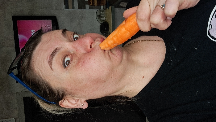 A medium close-up image depicts an adult female positioned indoors in Bel'kovo, Russia. She is looking directly forward with wide-open, brown eyes. Her lips are pursed and pushed outward around the tip of an orange carrot, which she holds with her right hand. Dark-rimmed eyeglasses with blue arms are perched on her head, above her hairline. She wears a dark t-shirt, a thin gold-colored chain necklace, and a ring with a light-colored stone on the fourth finger of her right hand. The background consists of a dark gray, textured wall. To the upper right, a television screen with a dark bezel displays a pink and magenta image with a partial white letter "M." Indistinct objects, possibly glass containers, are visible on a surface to the right.