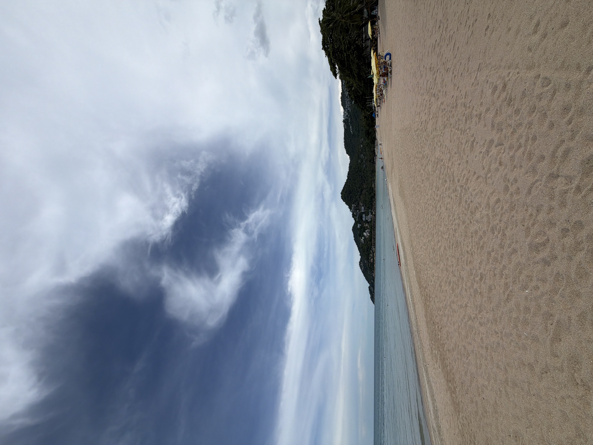 The image depicts a wide, sandy beach in Baan Chaweng, Thailand. The ocean is calm with gentle waves lapping the shore. In the distance, a lush green, hilly landscape dotted with buildings rises from the coast. The sky is partly cloudy with dramatic formations of dark and light clouds. Along the right side of the beach, a row of yellow umbrellas shades lounge chairs, some of which are occupied by people. Further down the beach, several individuals are visible, some near the water's edge and others near the umbrellas. A red object, possibly a surfboard or kayak, rests on the sand near the water. The overall scene conveys a relaxed, tropical atmosphere on a somewhat overcast day.
