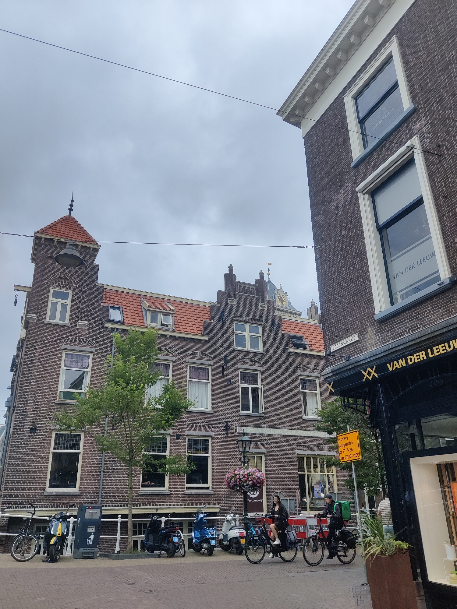 In the city of Delft, Netherlands, under an overcast sky, a street scene unfolds. Two individuals are depicted on bicycles, one wearing a green delivery backpack, suggesting a delivery service. They are cycling past a brick building with a red-tiled roof, adorned with multiple windows. Parked in front of the building are several scooters and a bicycle. A sign on the right side of the image reads "KOORNMARKT" and another sign is visible with text in Dutch that includes "DEZE WEG IS AFGESLOTEN". The foreground shows a brick-paved street.