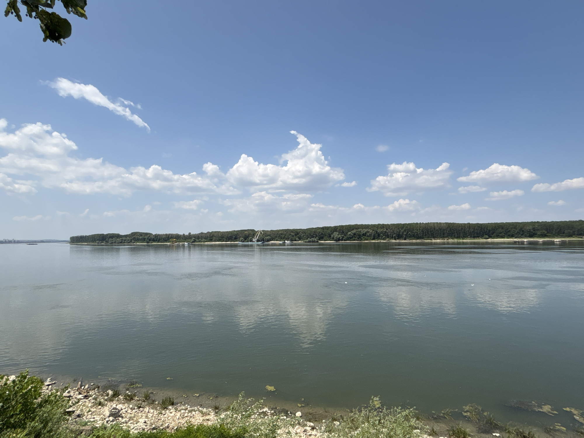 A wide, placid river flows under a bright blue sky dotted with fluffy white clouds. The riverbanks are lined with dense, green trees, and on the far bank, a sandy shore is visible with a few boats and a large crane. The water reflects the sky and clouds, creating a serene and peaceful scene. In the foreground, a rocky embankment is covered with sparse green vegetation. The lighting suggests it is a sunny day, likely in the late morning or early afternoon. The overall mood is tranquil and natural. There is no visible text in the image.