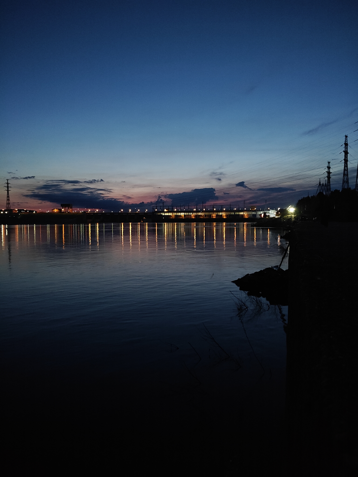 The photograph captures a wide-angle view of a river scene at twilight, likely in Chaikovsky, Russia. The sky dominates the upper portion of the frame, displaying a deep indigo blue that subtly transitions to a lighter blue and hints of orange-pink closer to the horizon, where scattered dark clouds are visible.
On the distant bank of the river, a long, horizontally oriented structure, possibly a dam or a hydroelectric power station, stretches across the midground. This structure is brightly lit by numerous artificial lights, which cast striking, elongated reflections that ripple across the calm surface of the water. These reflections appear as vibrant vertical streaks of orange and yellow light, providing a strong focal point.
Several prominent power transmission towers are silhouetted against the sky. One is visible on the far left, while multiple towers with their characteristic latticework are clustered on the right side of the frame, extending upwards with a network of power lines. The foreground on the right consists of a dark, indistinct bank or shoreline, possibly rocky or earthen, with very faint suggestions of vegetation.
The scene conveys a sense of peaceful twilight, contrasting the natural beauty of the fading light with the significant human-made infrastructure. The water appears still, reflecting the distant lights with clarity. No people or visible text are present in the image. The setting suggests an industrialized area bordering a large body of water, consistent with the location context of Chaikovsky and its major river infrastructure.