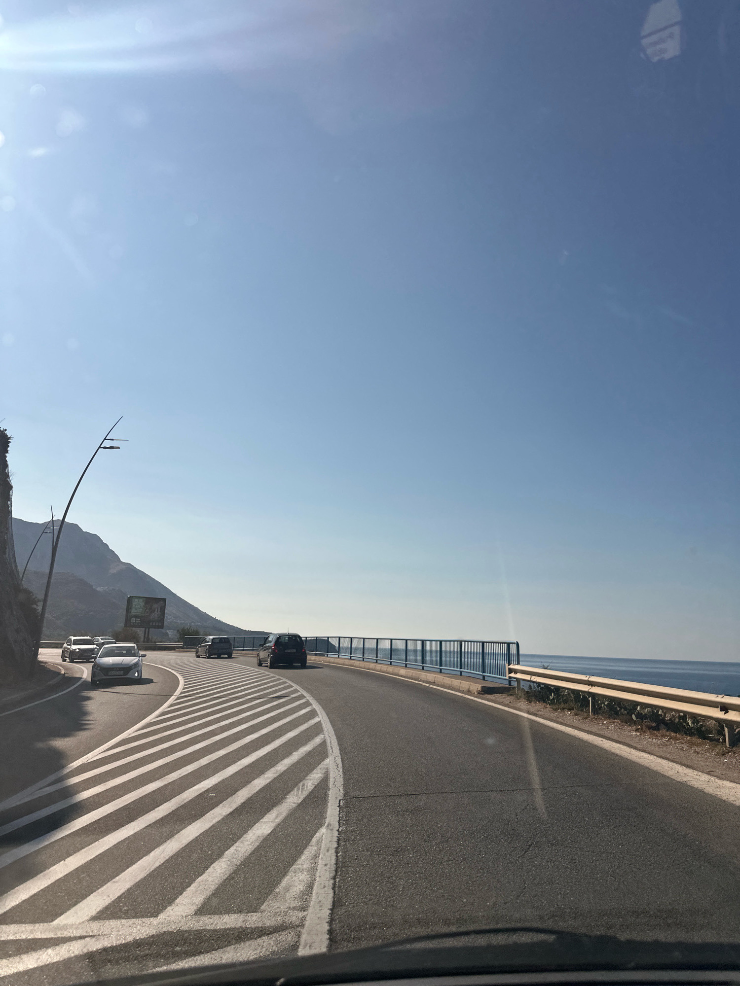 The image depicts a scenic coastal road in Rafailovići, Montenegro, on a bright, sunny day. The perspective is from inside a vehicle, looking forward and slightly to the left. The road curves to the right, with a distinct pattern of white diagonal stripes marking a traffic lane or a pedestrian crossing area. On the left side of the road, a steep, rocky hillside rises, topped with sparse vegetation and a tall, modern-style lamppost. A billboard is visible on the hillside.
Several cars are traveling on the road. In the foreground, a white car is in the lane closest to the viewer, followed by other vehicles. On the right side of the road, a blue metal railing separates the road from a drop-off towards the sea. Beyond the railing, the vast expanse of the blue Adriatic Sea stretches to the horizon. The sky is a clear, bright blue with a slight haze and lens flare from the sun, which is positioned in the upper left corner of the frame. The overall scene conveys a sense of travel along a beautiful, mountainous coastal route.