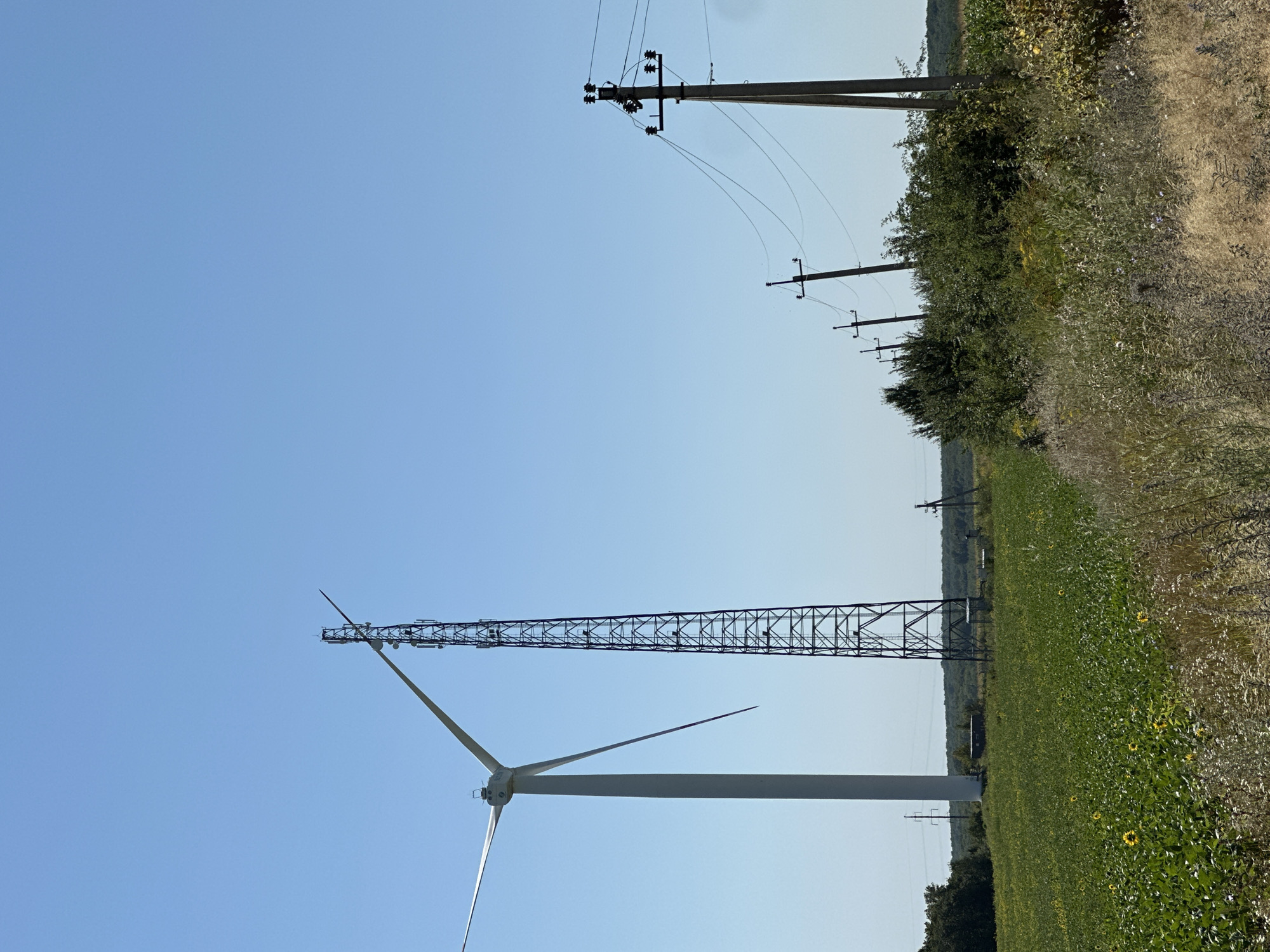 The image depicts a rural landscape with several tall structures against a clear blue sky. In the foreground, a vast green field is visible, with some scattered sunflowers. To the left, a white wind turbine stands tall, its blades partially visible. Next to it, a tall, dark, lattice-like tower, likely a cell tower or communication mast, rises high into the sky. Further to the right, a series of utility poles with electrical wires stretches across the frame, leading towards a distant line of trees. The overall scene suggests an open, possibly agricultural, area in the countryside. The location is specified as Korchivka, Ukraine. There are no people visible in the image.