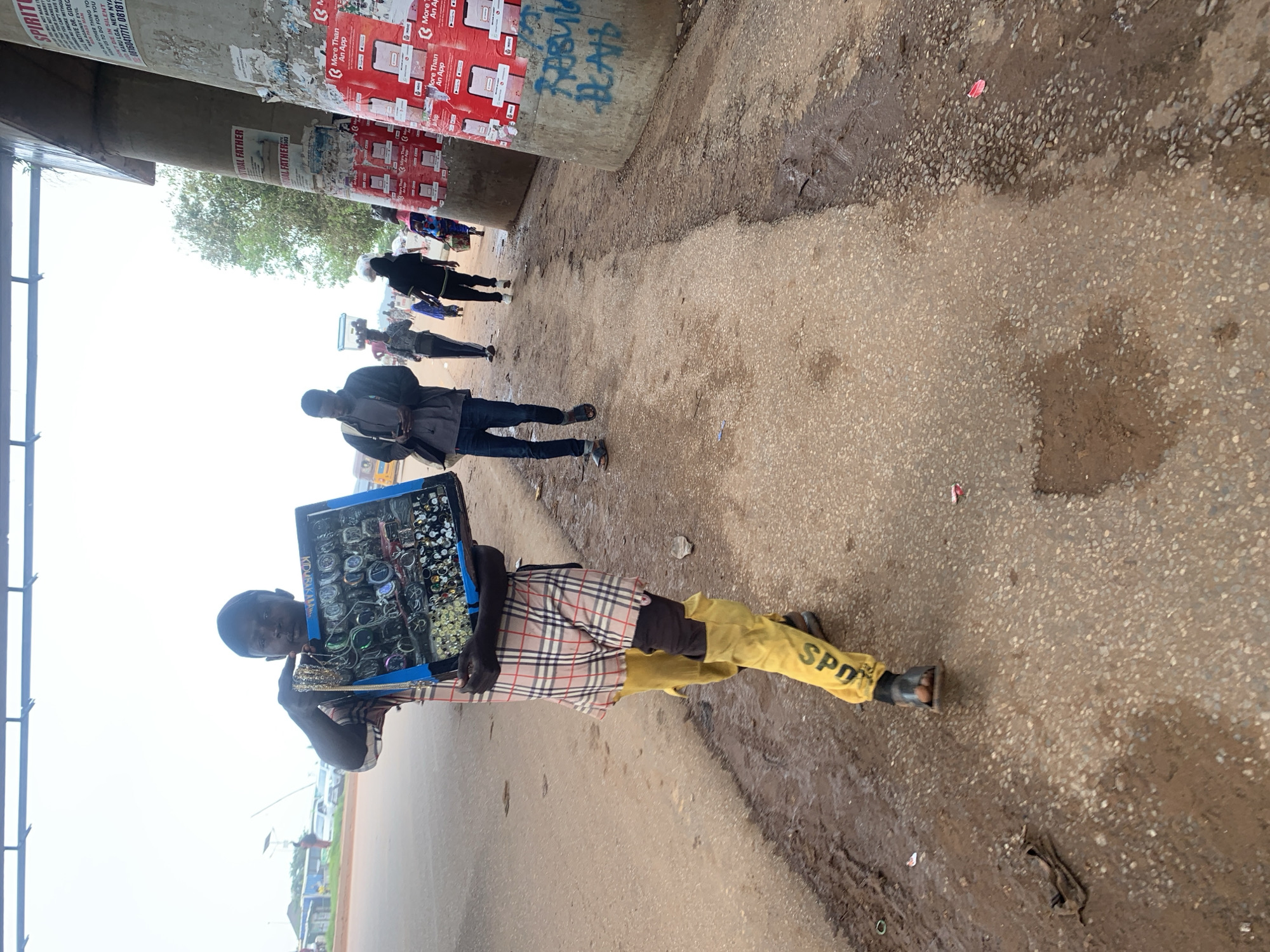 In Nyanya, Nigeria, a young man walks along a paved road, holding a display box of jewelry and watches. He wears a plaid shirt and bright yellow pants with the word "SPORT" printed on them. Behind him, other people are walking, some carrying items on their heads. The scene takes place under a concrete overpass, where advertisements and graffiti adorn the pillars. The road is wet and muddy in places, indicating recent rain.