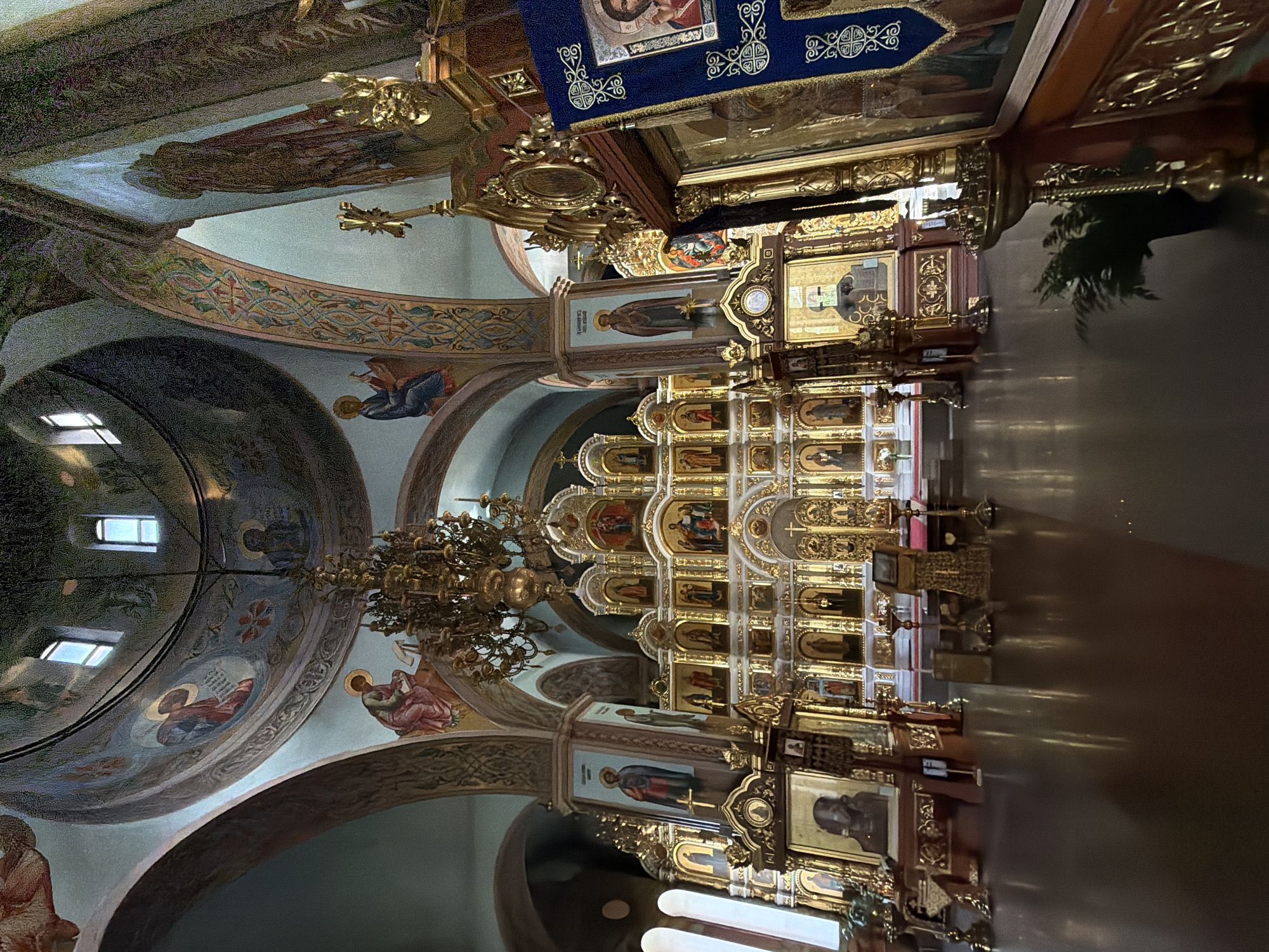 The image depicts the interior of the Ascension of the Lord Orthodox Church in Chaikovsky, Russia. The scene is dominated by the ornate iconostasis, a wall of religious icons that separates the nave from the sanctuary. The iconostasis is richly decorated with gold leaf and features numerous framed religious paintings, known as icons. To the left of the iconostasis, a large, elaborate chandelier hangs from the domed ceiling, which is adorned with frescoes of religious figures.
The environment is dimly lit, with the primary light sources appearing to be the windows in the dome and the golden glow emanating from the iconostasis and other decorative elements. The floor is highly polished, reflecting the light and the architectural features of the church.
In the foreground, on the right side of the image, a large icon is displayed on a wooden stand, draped with a blue cloth featuring decorative patterns. Several ornate brass candle holders are visible throughout the space. A potted plant is also present near the foreground.
No people are visible in the image. The overall impression is one of solemnity and grandeur, characteristic of Eastern Orthodox church interiors. The architectural style suggests a traditional design with elements of Byzantine influence.