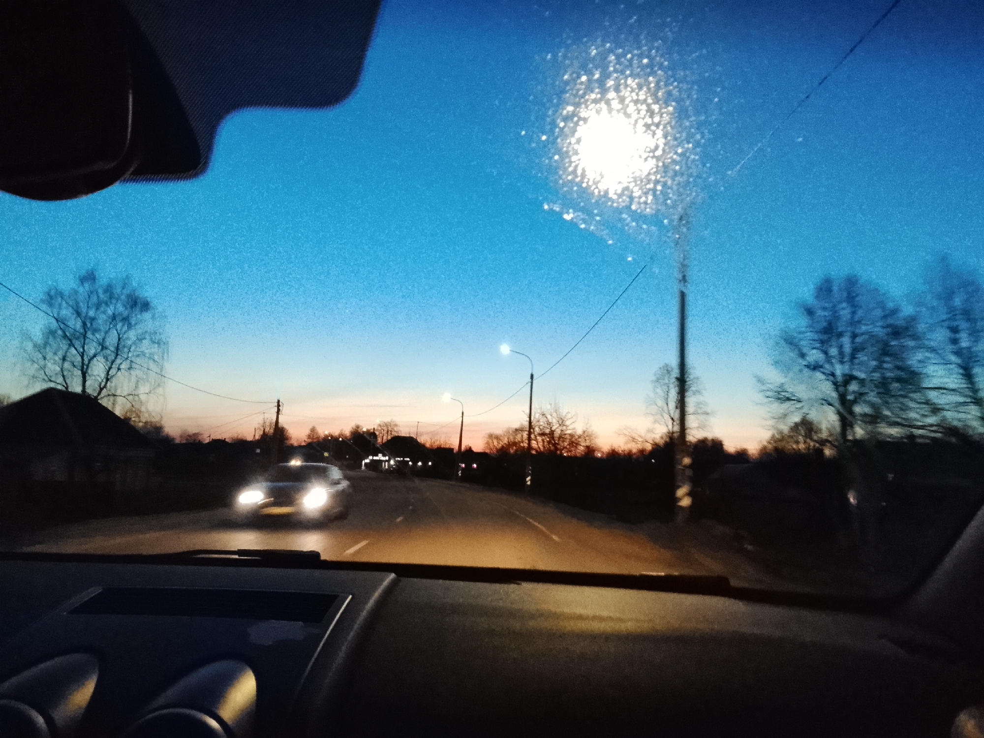 The image shows a view from inside a car, looking through the windshield. It is dusk or early evening. A road stretches ahead, illuminated by car headlights and streetlights. Another car with headlights on is approaching in the opposite direction. Bare trees are visible on either side of the road. The sky is a gradient of blue to orange. The dashboard of the car is in the foreground. The location is likely Высокиничи, Russia, based on the prompt.