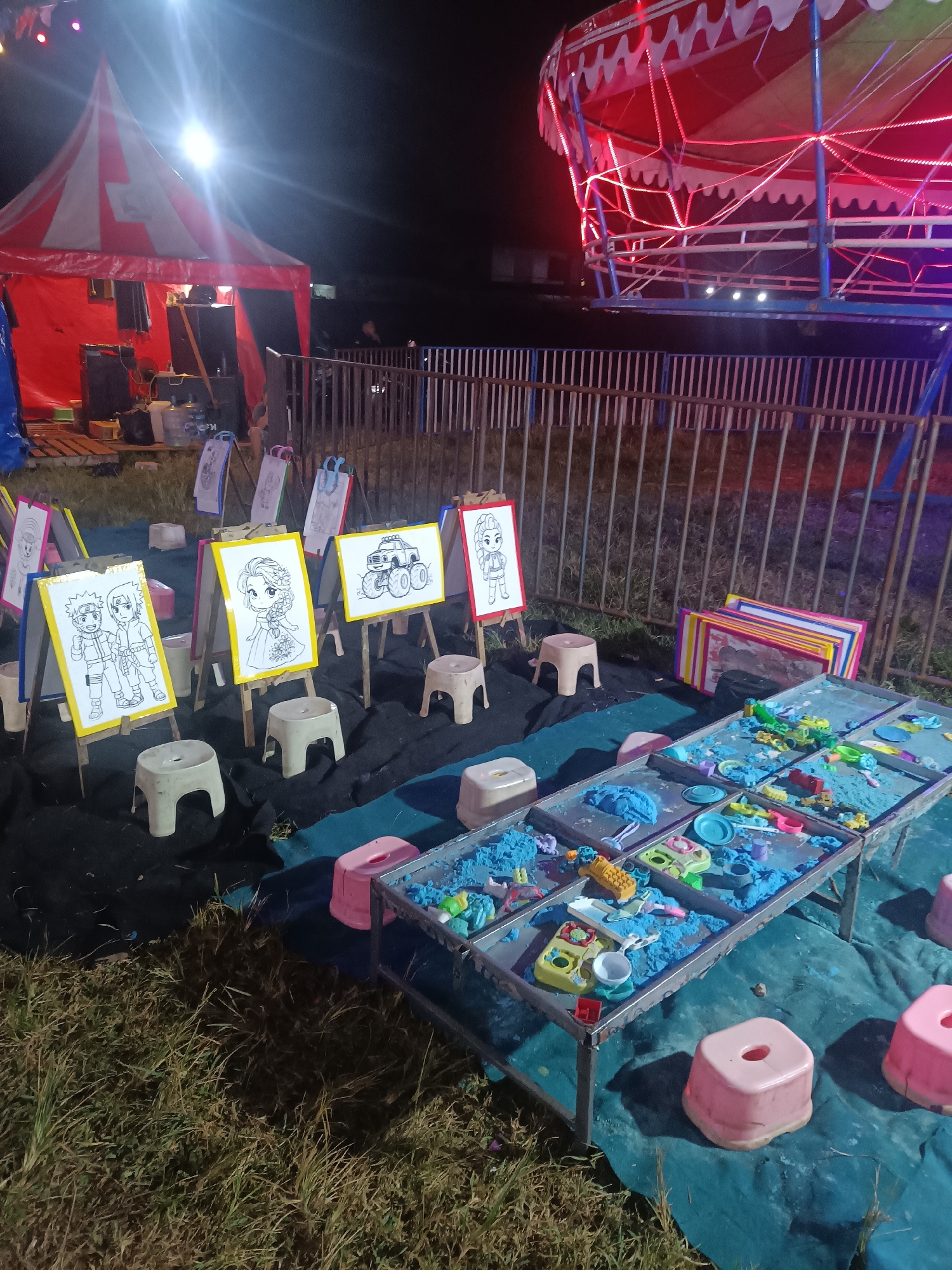 The image depicts an outdoor nighttime scene at what appears to be a carnival or fair, likely in Depok, Indonesia. The foreground is dominated by a play area with tables filled with blue kinetic sand and various plastic toys, suggesting children's activities. Several small, light-colored stools are scattered around the sand tables and in front of easels displaying coloring pages.
Behind the play area, a line of easels stands, each holding a coloring page featuring cartoon characters and a monster truck. Some of these easels have bright, colored borders. A metal fence runs across the mid-ground, separating the activity area from other attractions.
In the background, a large tent with red and white stripes is visible, illuminated by bright lights, suggesting a performance or vendor area. To the right, a colorful carnival ride, possibly a carousel or a similar spinning attraction, is lit up with red and white lights. The overall atmosphere is festive and inviting, with the artificial lighting creating a vibrant, albeit somewhat hazy, ambiance. The presence of grass in the foreground indicates an outdoor setting. No people are clearly discernible in the image, but the setup implies a public event catering to families. The time of day is clearly night, and there are no strong indicators of specific weather conditions.