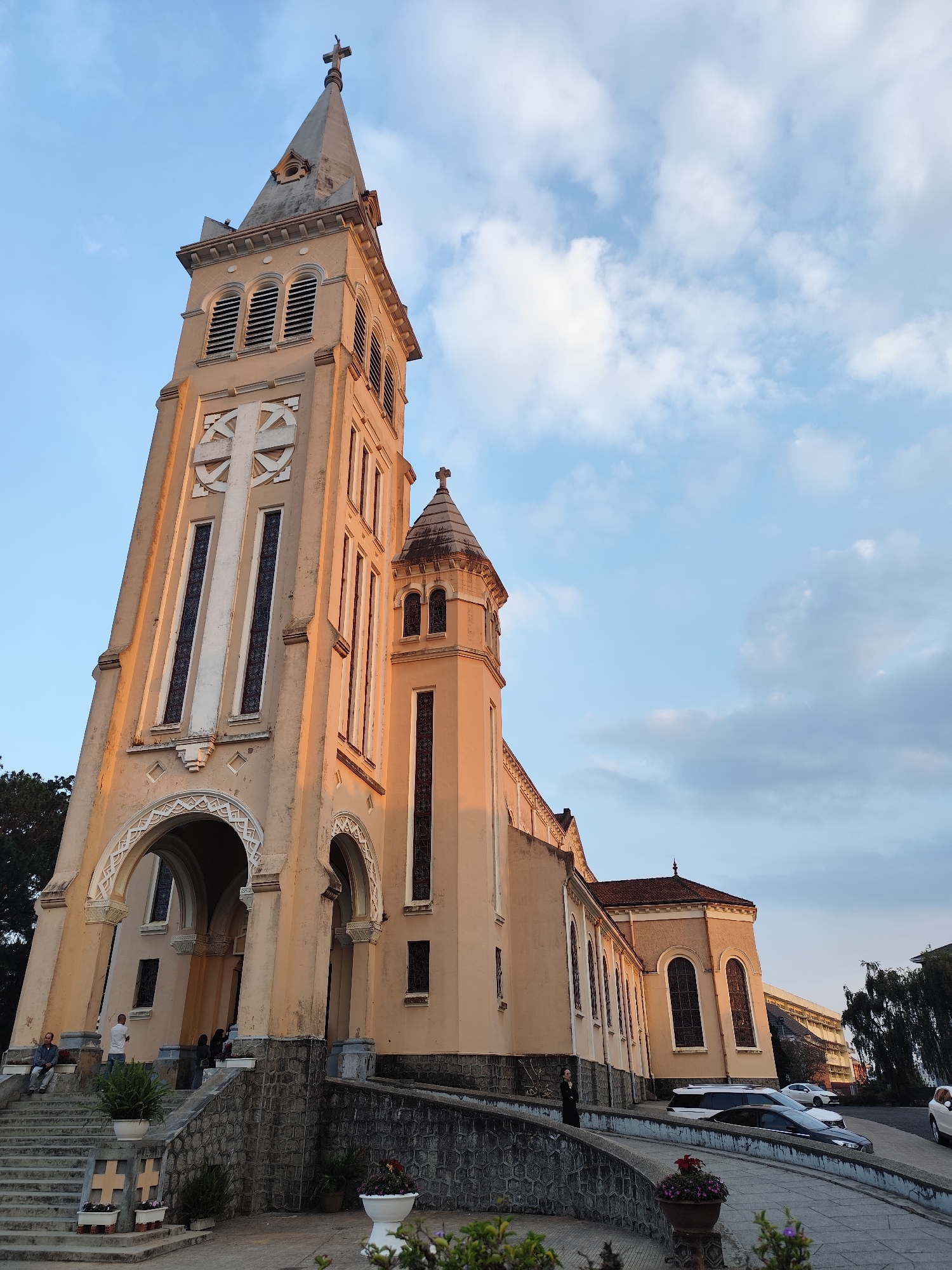 The image captures St. Nicholas Cathedral, a Roman Catholic cathedral located in Da Lat, Vietnam. The imposing structure is made up of warm-colored stone, with a tall bell tower topped with a cross. Architectural details such as arches, ornate windows, and crosses adorn the façade. The cathedral is positioned on an elevated platform with stone stairs leading up to the entrance. People are visible on the steps and in the background. Cars are parked along the side of the building. The sky is visible, filled with clouds.