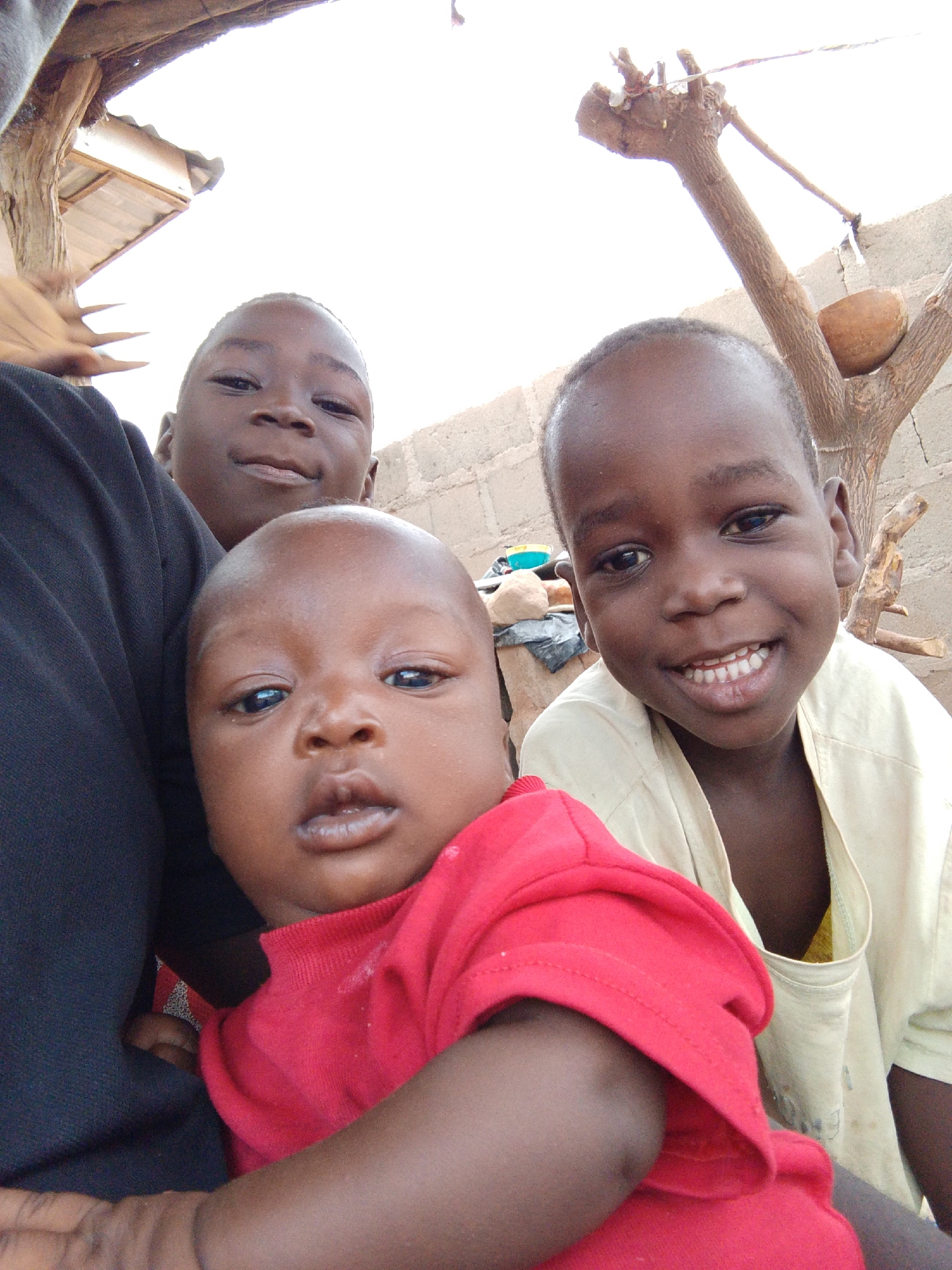 Three young children are clustered together in an outdoor setting in Mulke Barikin Sayejo, Nigeria. A baby wearing a red shirt is in the foreground. Behind the baby is a child wearing a black garment and another child wearing a light-yellow shirt. A tree branch with a bowl on it is visible in the background, along with a section of a concrete block wall. A partial view of a corrugated metal structure is above and to the left.